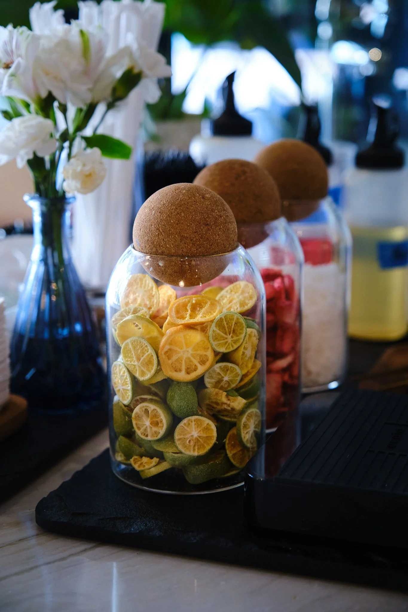A clear glass jar filled with slices of lemon and lime, topped with a round brown cookie, placed on a black surface with blurred background of flowers and bottles.