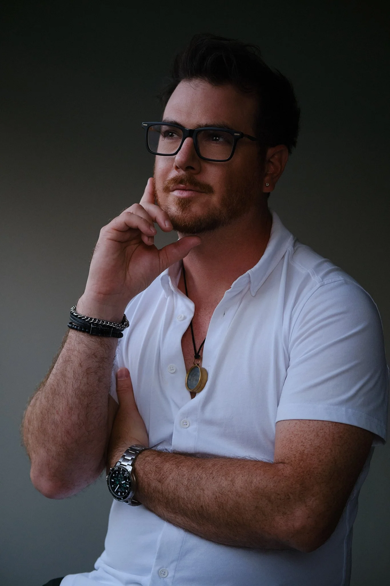A man wearing black glasses, a white button-up shirt, a wristwatch, and bracelets, is posing with his hand on his chin against a dark background.