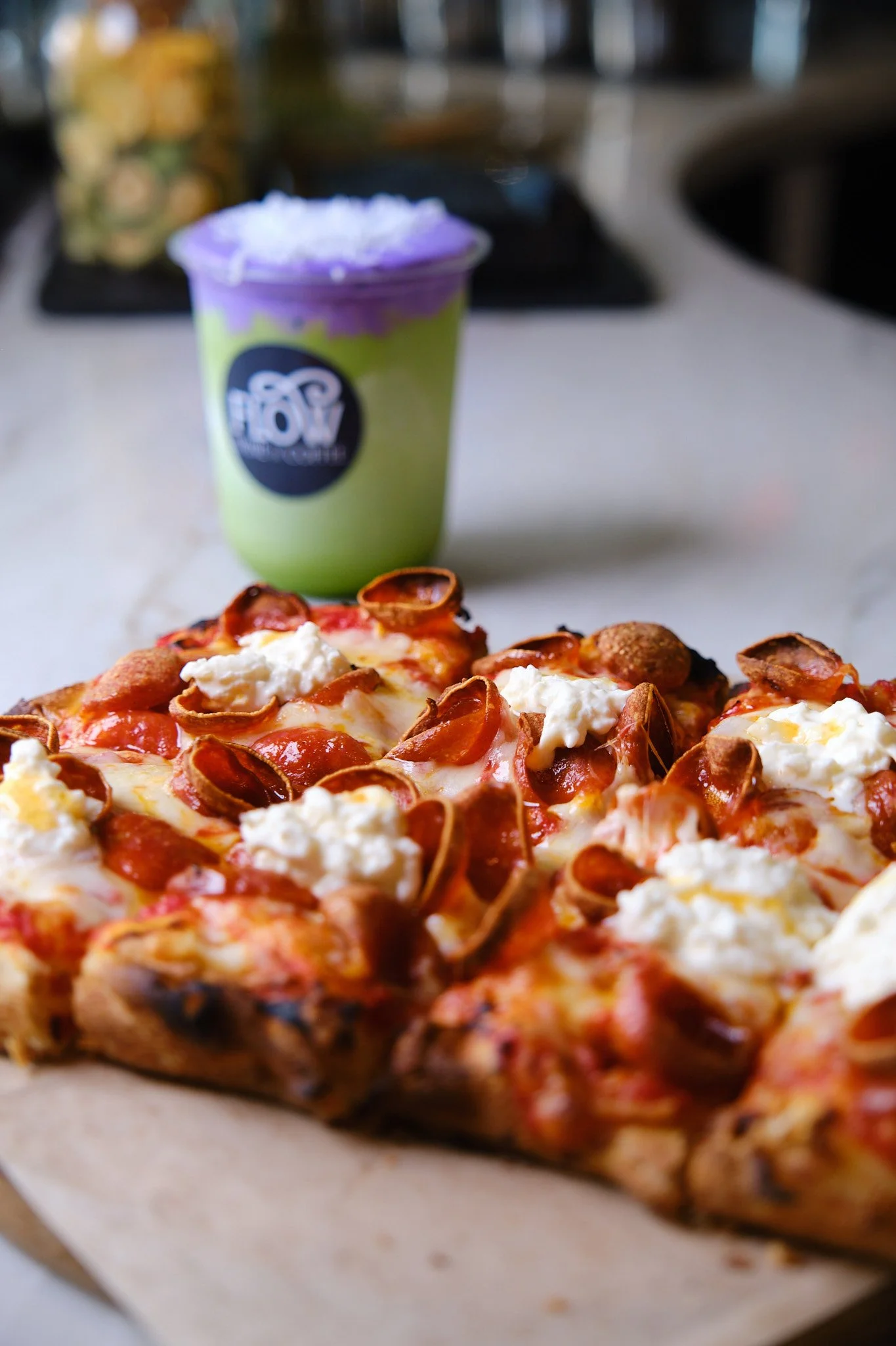 Close-up of a pepperoni pizza with dollops of ricotta cheese, served on parchment paper, with a green and purple soft drink in a cup labeled "POP" in the background.