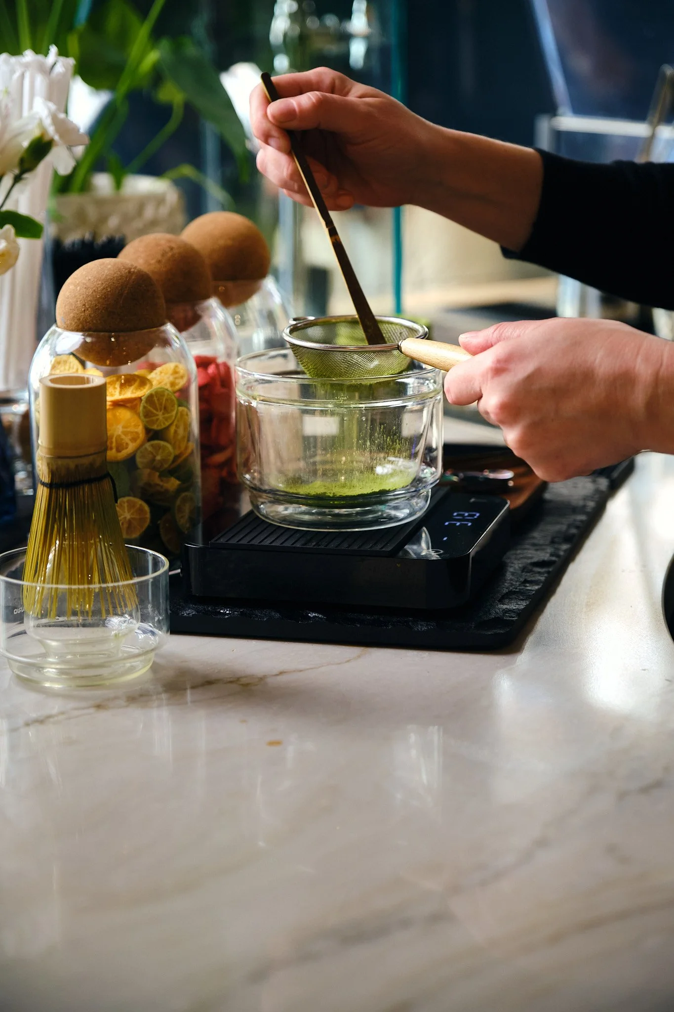 A person's hand holding a small sieve straining a green liquid into a glass bowl on a digital scale. There are jars of lemon slices and other ingredients on the counter, with plants and a dark background.