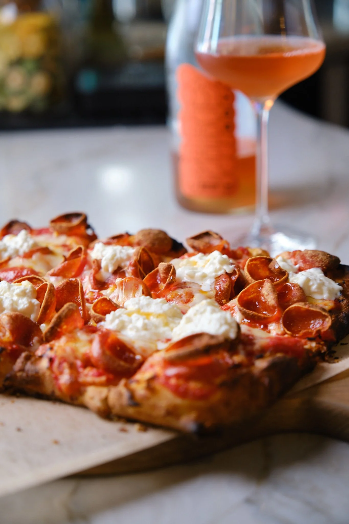 Close-up of a pizza with pepperoni and cheese on a baking sheet, with a glass of pinkish cocktail in the background