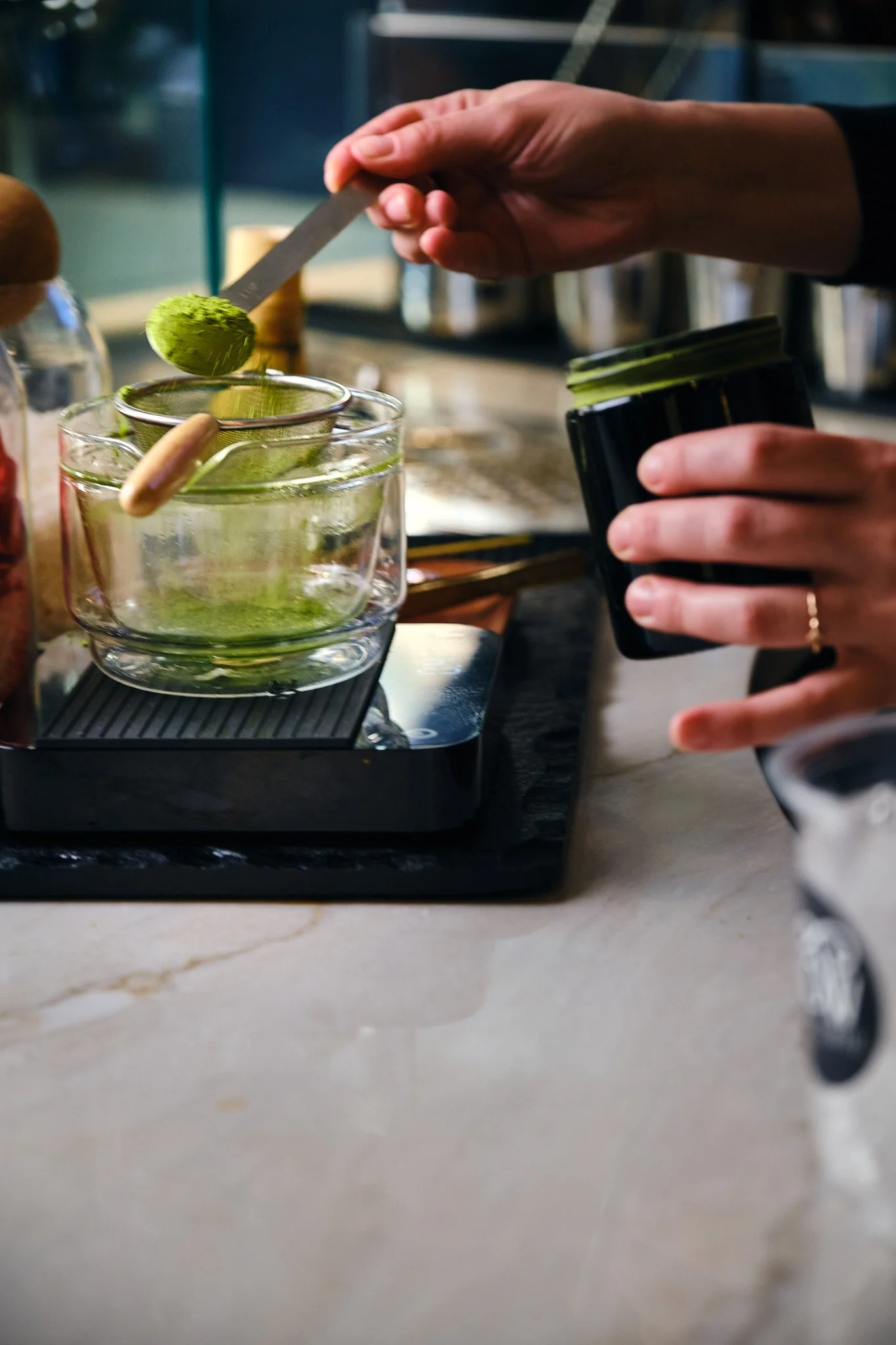 Person holding a small container of matcha powder over a glass cup with a metal strainer, preparing a matcha tea.