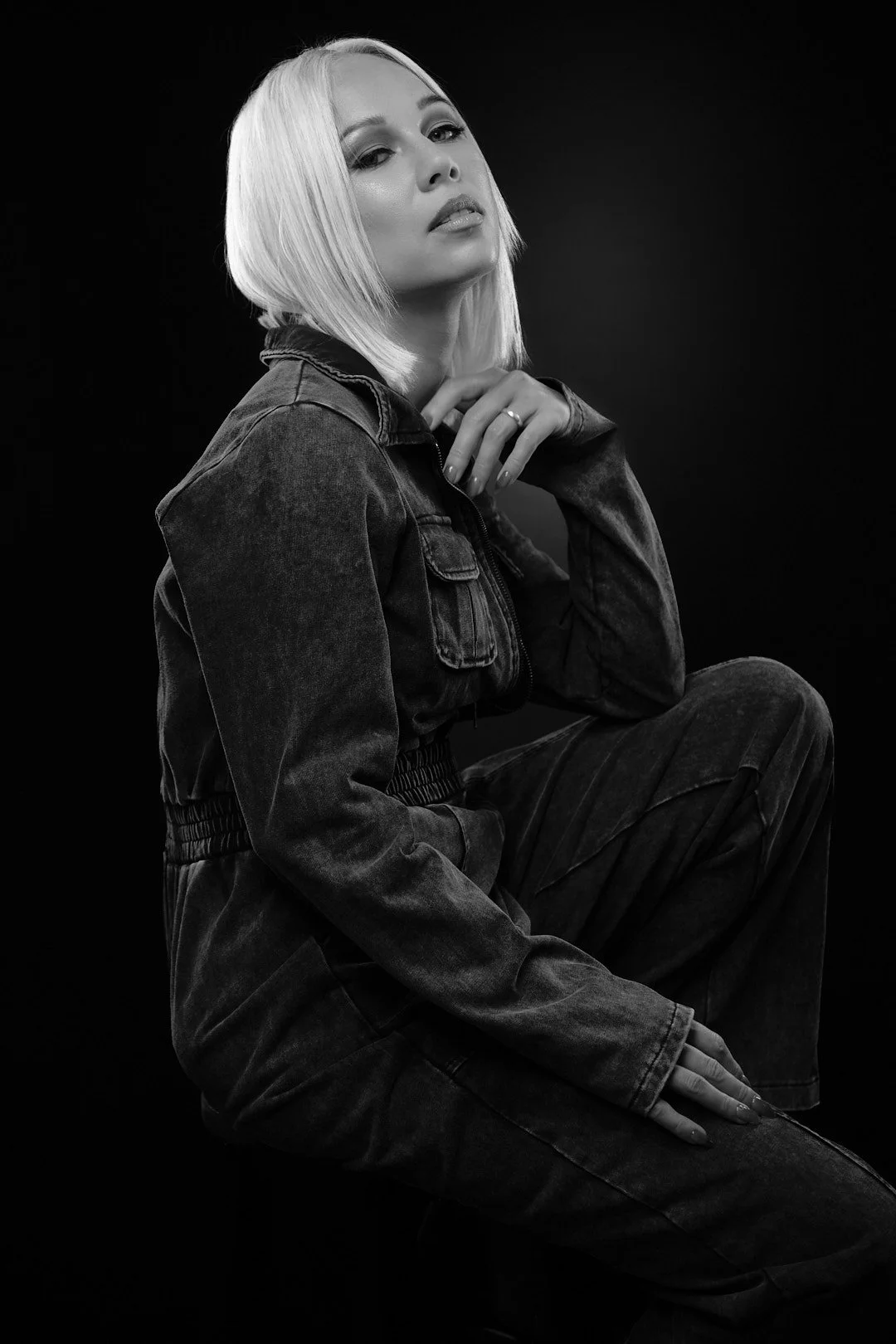 Black and white portrait of a woman with platinum blonde hair in a casual denim outfit, sitting against a dark background.
