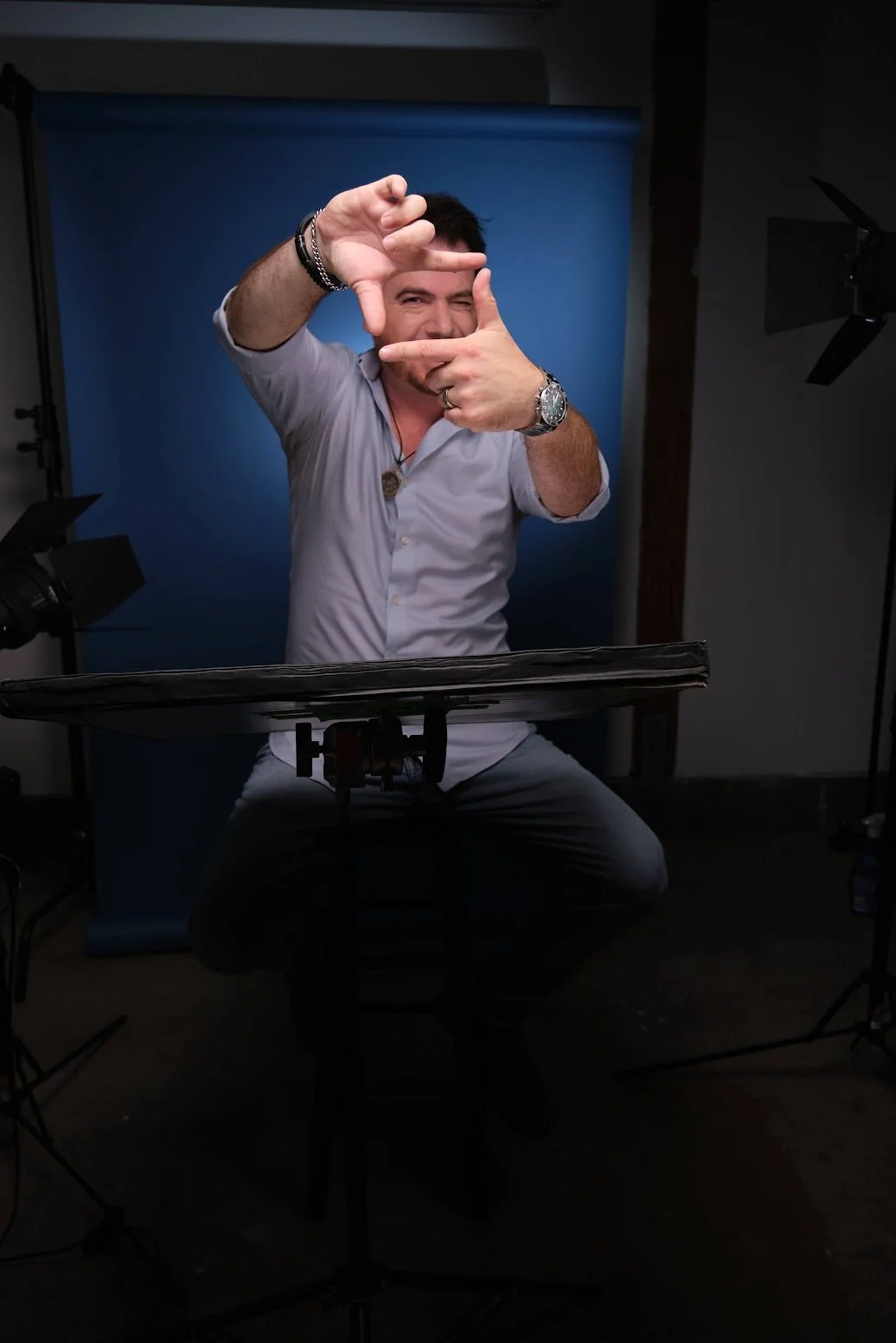 A man in a white shirt posing with his hands forming a frame, sitting at a table with photography studio equipment around him, with a blue backdrop behind him.