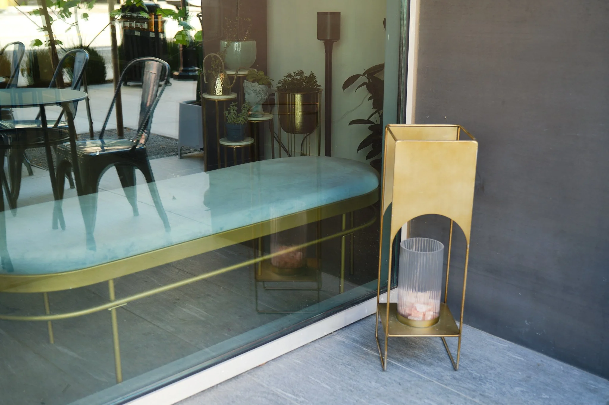 View of a stylish interior space through a glass window, showing a mustard-colored metal frame side table with a pink marble top, several potted plants on a white round stand, and black chairs around a round table outside. The scene includes reflecti