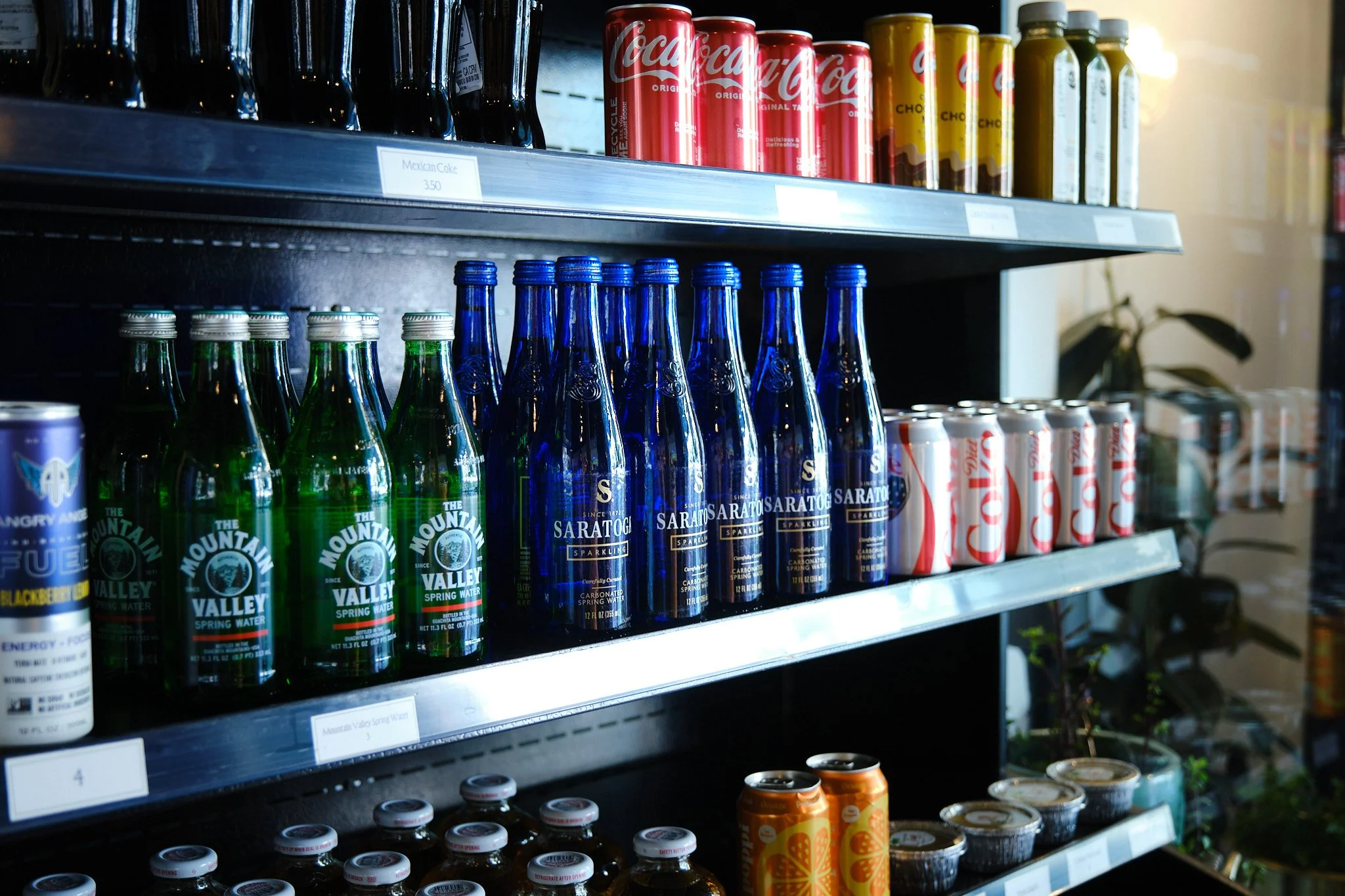 Shelves with bottled beverages including Coca-Cola cans, Saratoga sparkling water bottles in blue, Mountain Valley spring water in green bottles, and other assorted drinks in cans and bottles.