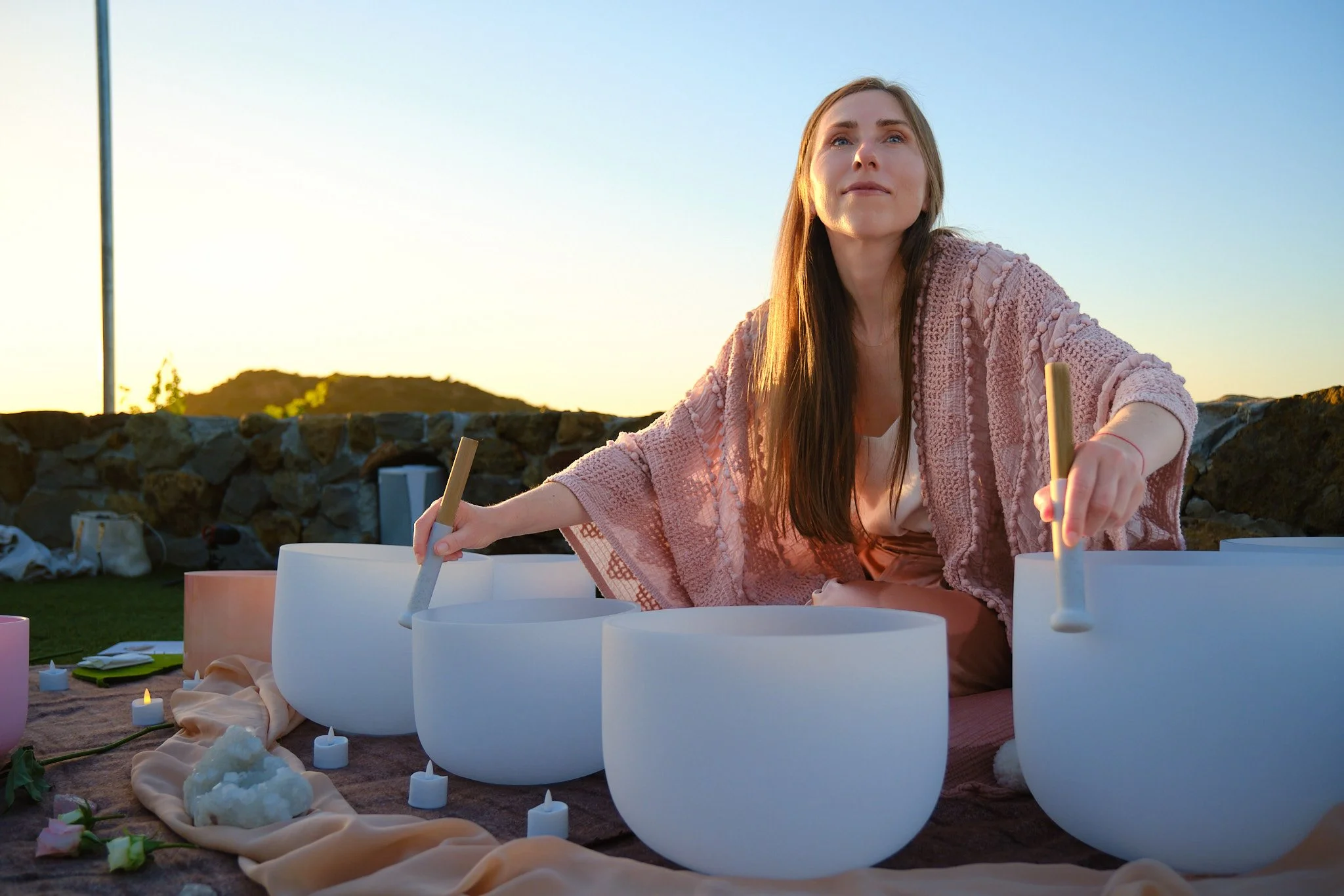 A woman playing singing bowls outdoors during sunset, surrounded by candles and soft fabrics.