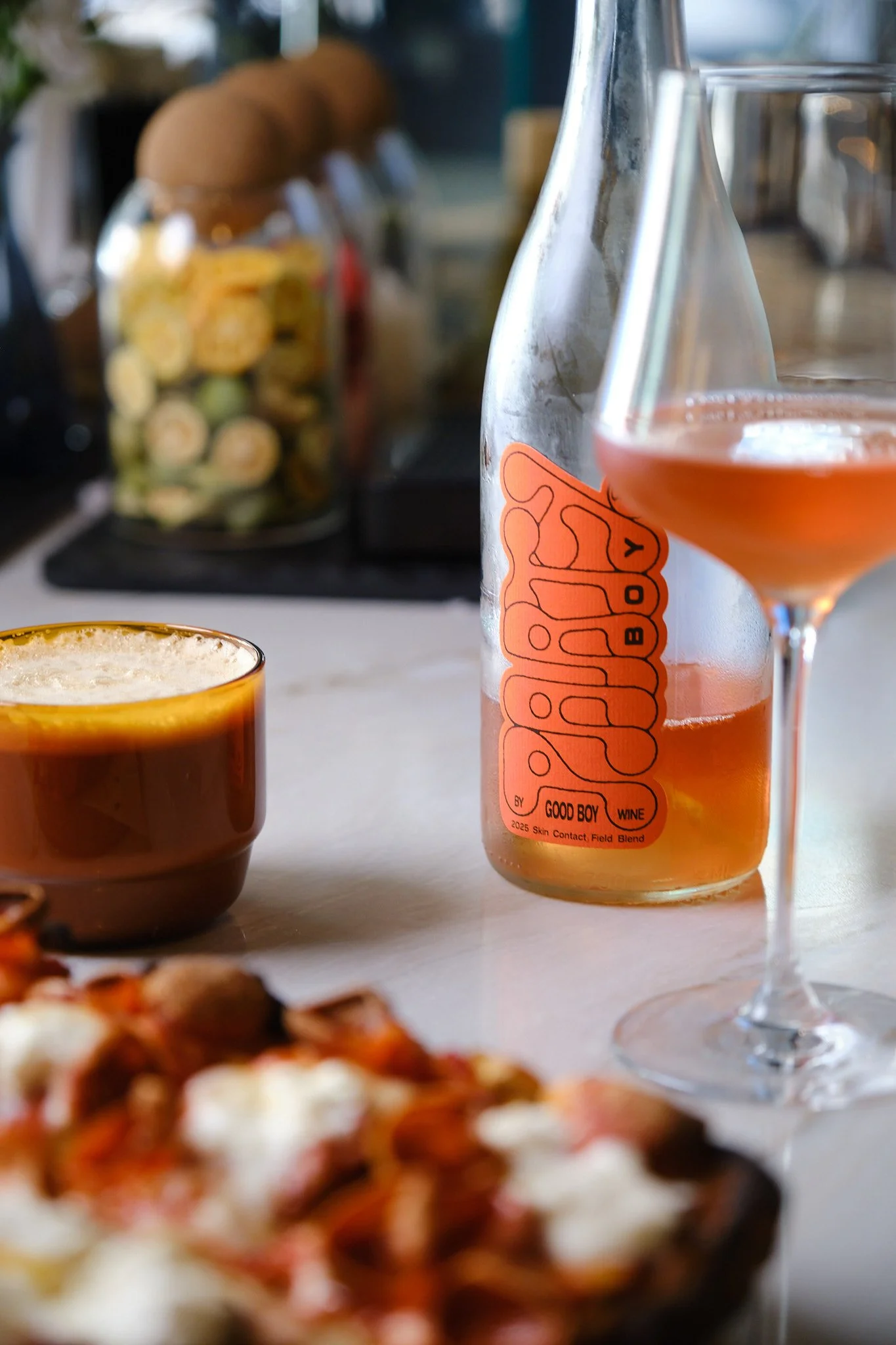Close-up of a glass bottle with orange label and a glass of pink wine in the foreground, with a jar of cookies and various kitchen items blurred in the background.