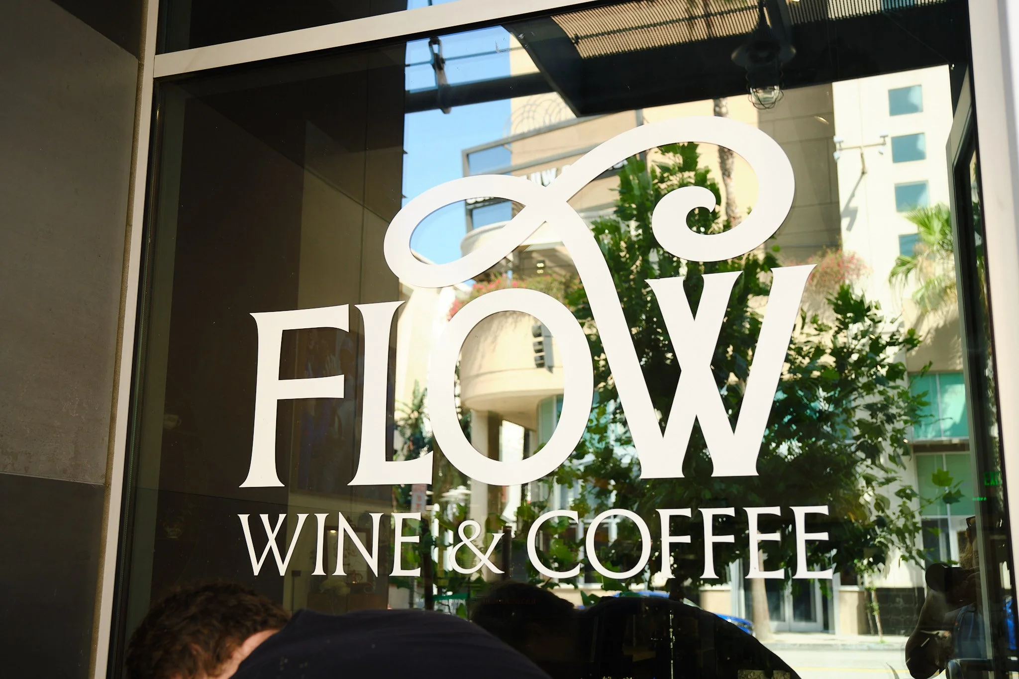 Store window with white text reading 'EQUINOX' and additional text indicating it sells wine and coffee, reflecting urban surroundings with buildings and greenery visible outside.