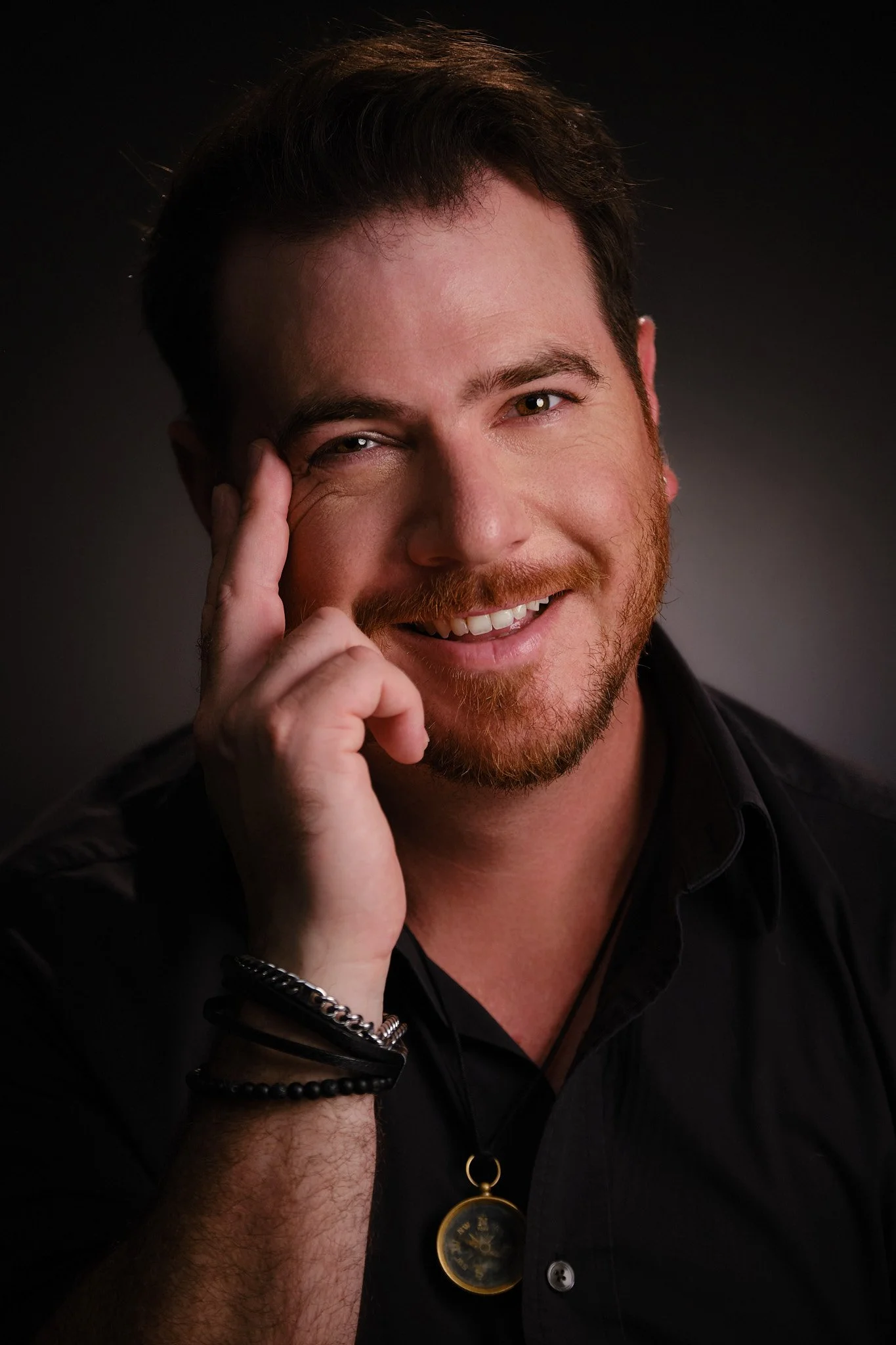 Close-up portrait of a smiling man with brown hair, a beard, wearing a black shirt, a necklace with a clock pendant, and bracelets, posing against a dark background.