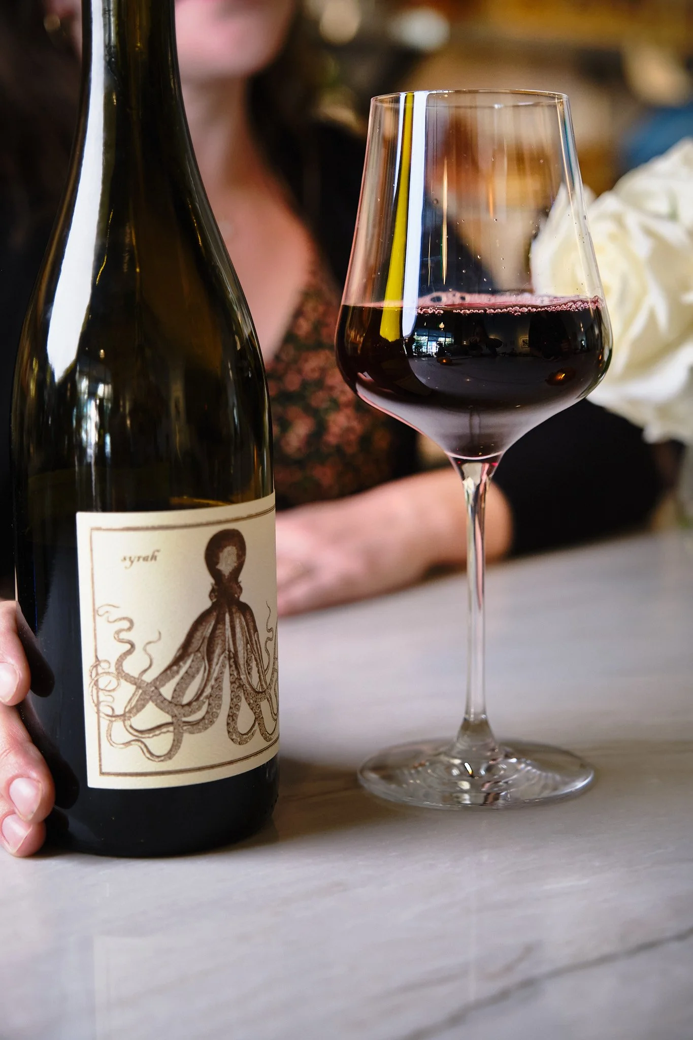 A person holding a bottle of red wine next to a filled wine glass on a white table. The label on the bottle features an octopus illustration.