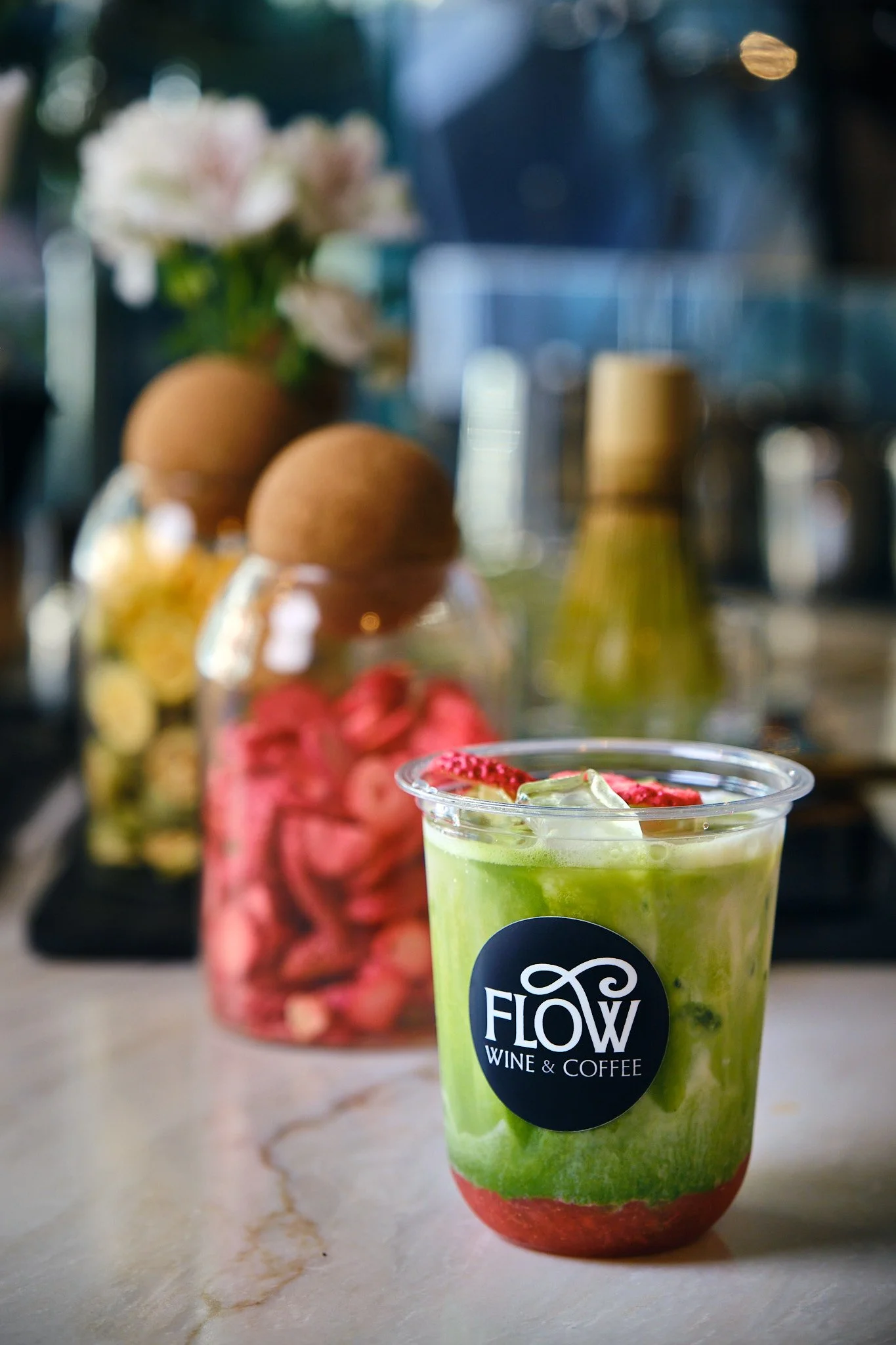 A green smoothie in a clear plastic cup with a black label reading "Flow Wine & Coffee" in the foreground, with a jar of pink pretzels, a jar of red candies, two brown eggs, and a blurred background of flowers and kitchen objects.