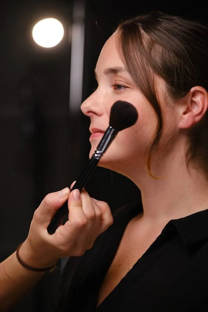 A woman with brown hair getting her makeup applied with a black makeup brush by a person with a brown bracelet, against a dark background with a bright round light.