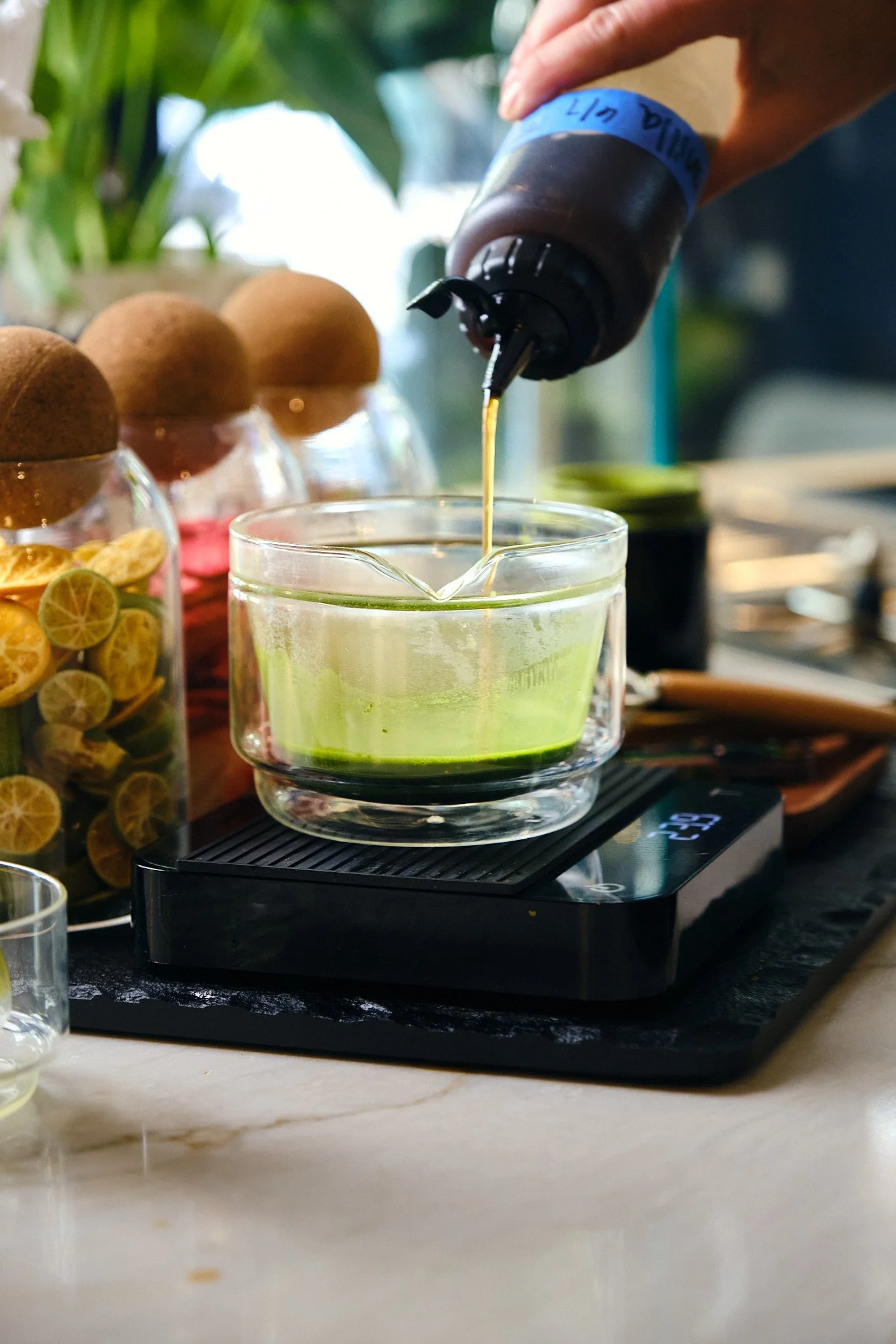 A hand pouring a dark liquid from a squeeze bottle into a glass container with a yellow-green liquid, on a digital scale. Three brown eggs on egg cups are in the background, along with a glass jar of sliced lemon and lime slices. Green plant leaves a