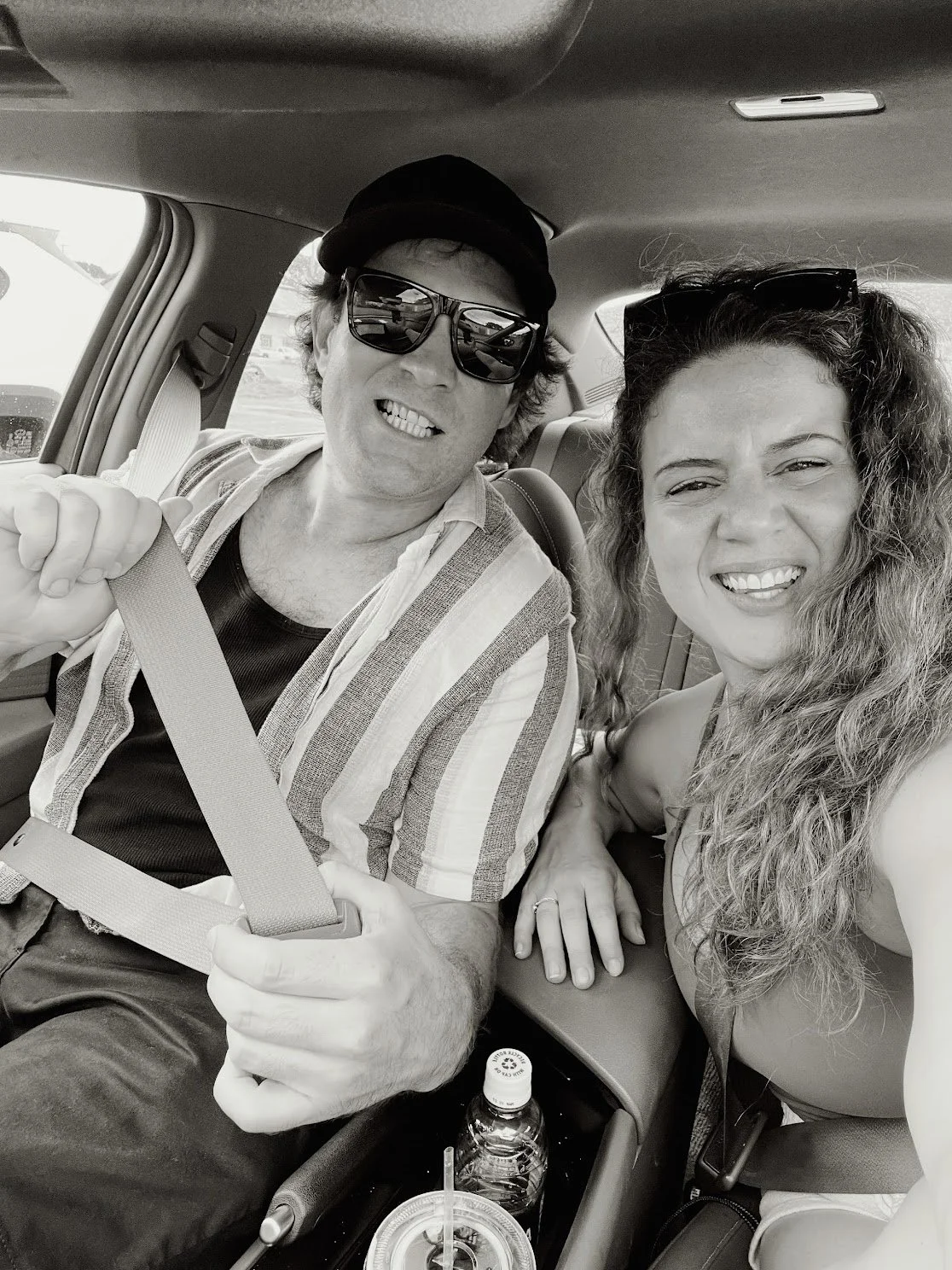 A black and white photo of a man and woman sitting in the front seats of a car, smiling at the camera. The man is wearing sunglasses and a striped shirt, holding a seatbelt. The woman has long curly hair and sunglasses on her head.