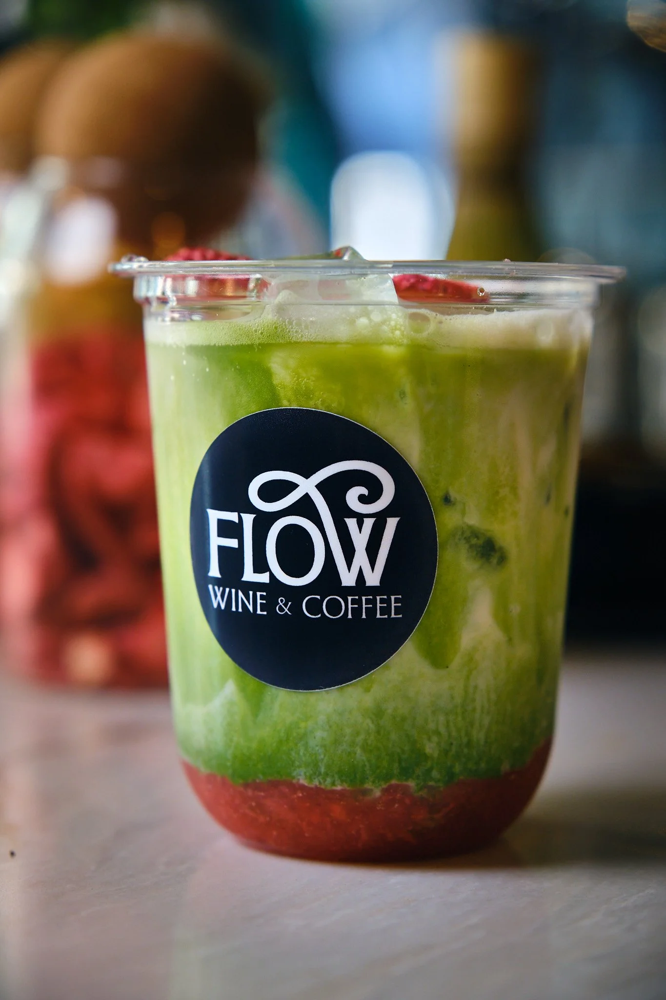 Close-up of a green smoothie or juice in a clear plastic cup with a black circular logo that reads 'Flow Wine & Coffee'. The background shows a blurred red fruit, possibly strawberries, and other colorful blurred objects.