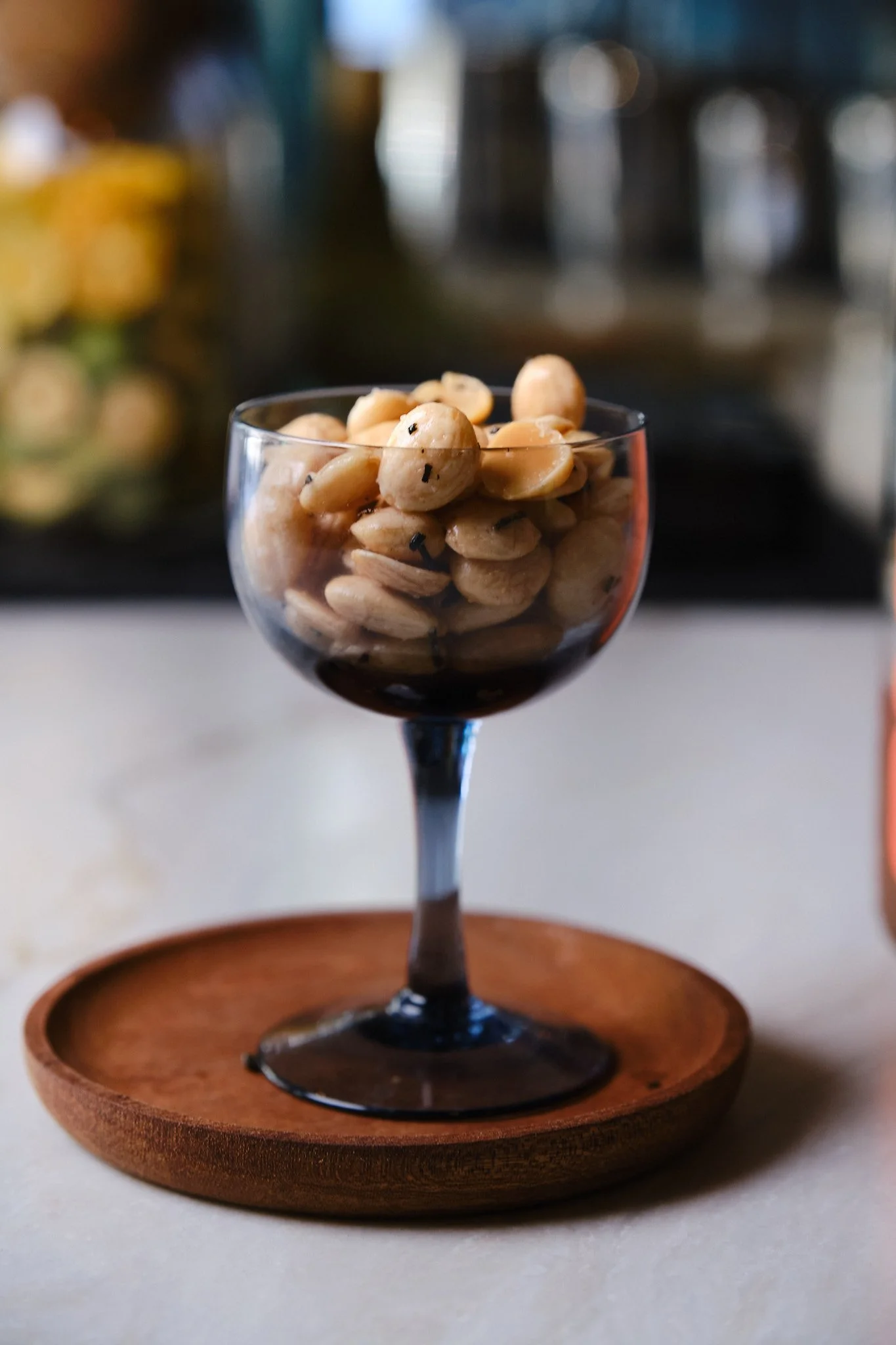 A glass of mixed nuts with black sesame seeds on top, placed on a wooden coaster.