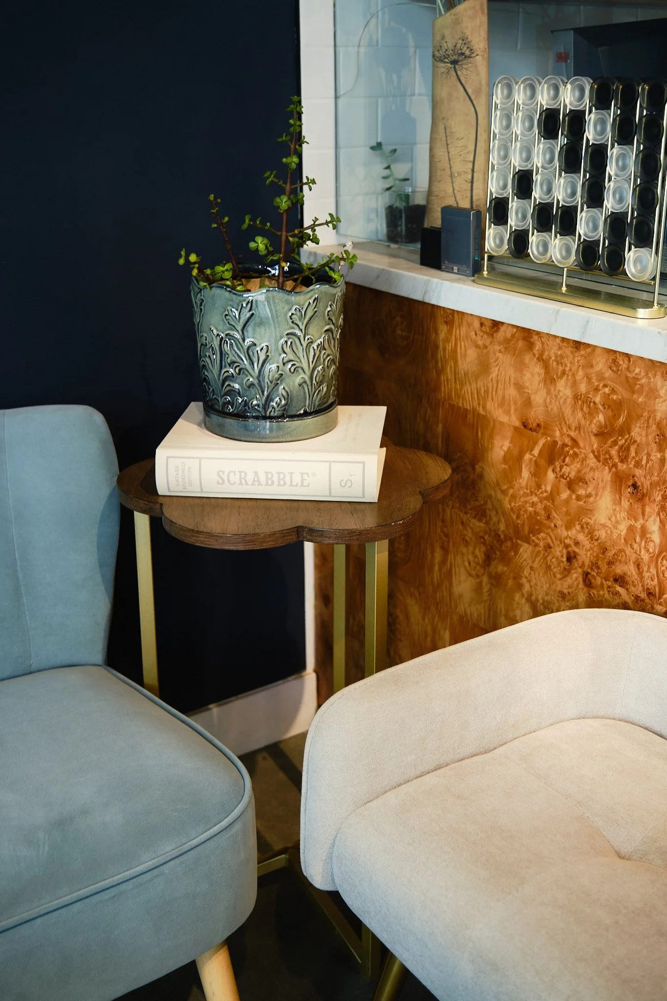 Interior view featuring a table with a ceramic pot of green succulent, a book titled 'SCRABBLE', light-colored upholstered chairs, a wooden counter, and decorative items including a dandelion drawing, a small black speaker, and a metal spice rack.