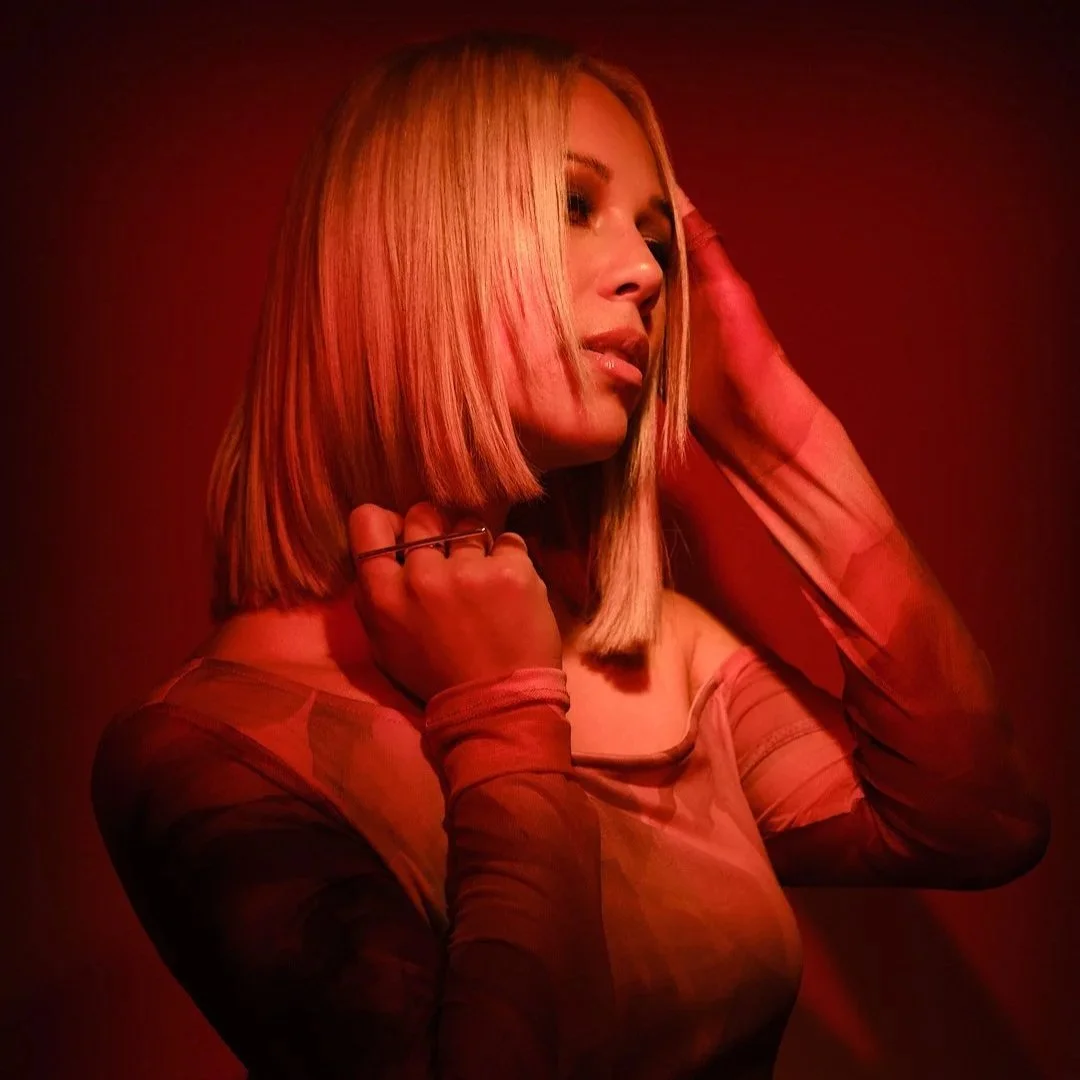 A woman with blonde hair in a bob hairstyle posing against a red background. She is wearing a sheer, long-sleeved top and has a contemplative expression, with her hand near her head.