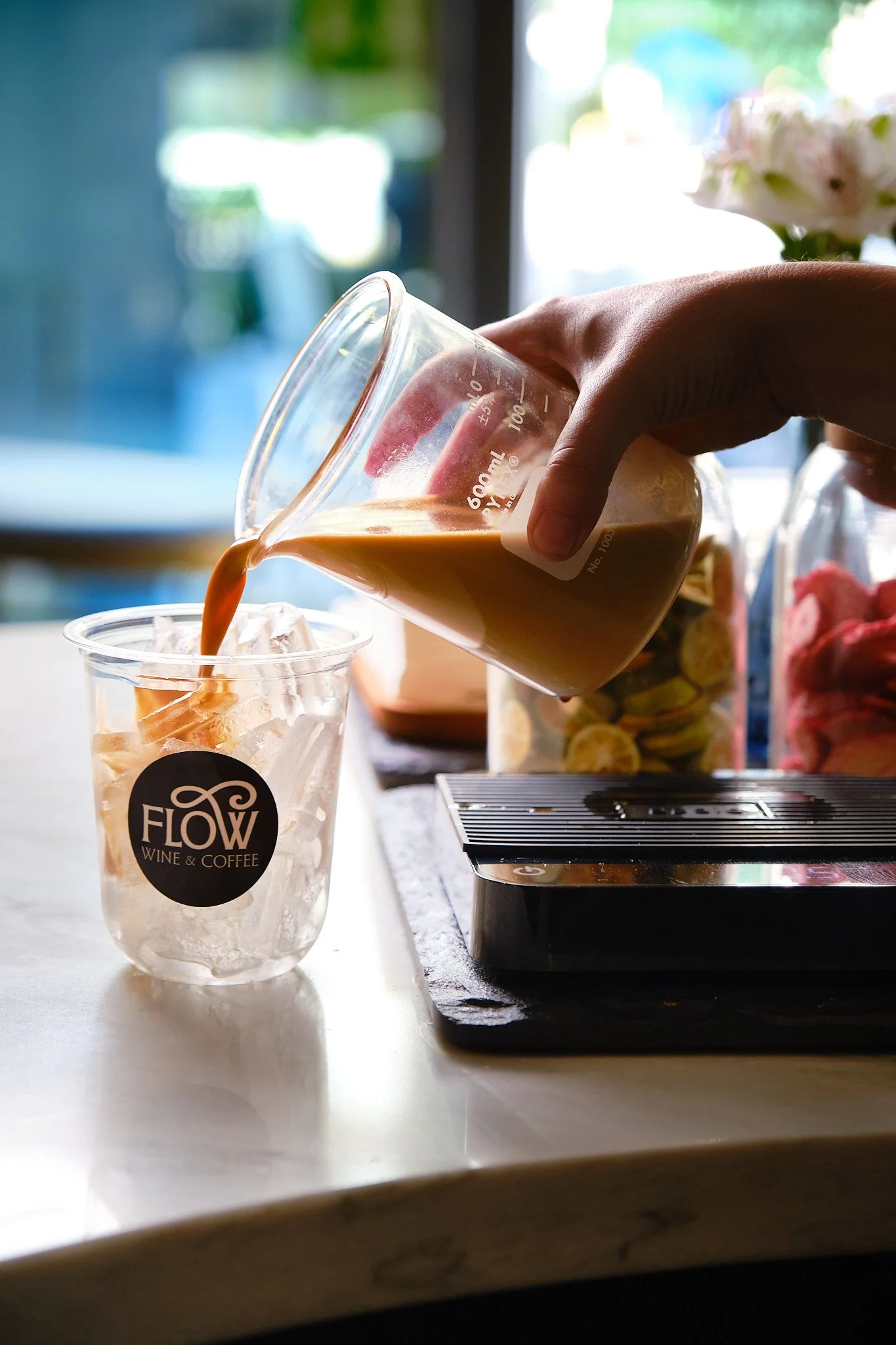 A person pouring coffee into a glass with ice, located on a white surface near a black scale. In the background, there are bottles with colorful candies, a flower, and a window with blurred green scenery outside.