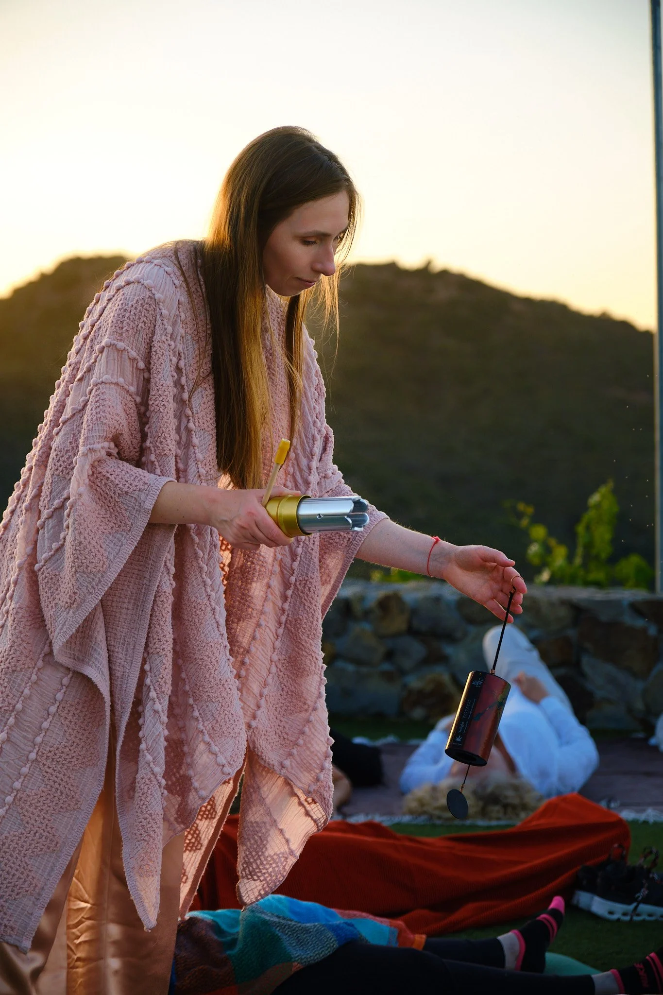 A woman with long brown hair wearing a pink textured shawl and tan pants standing outdoors during sunset, holding a cylindrical device with a yellow top in her right hand, with mountains in the background.