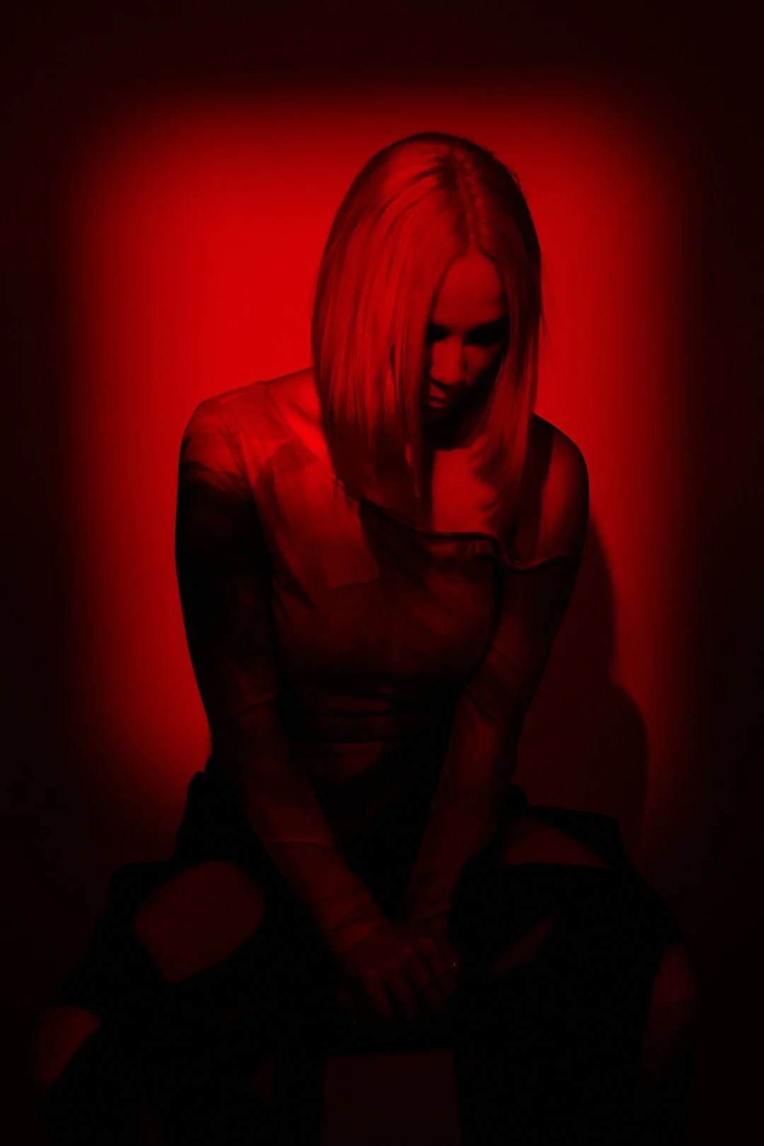 A woman with blonde hair sitting against a red background, with her head bowed and looking downward.