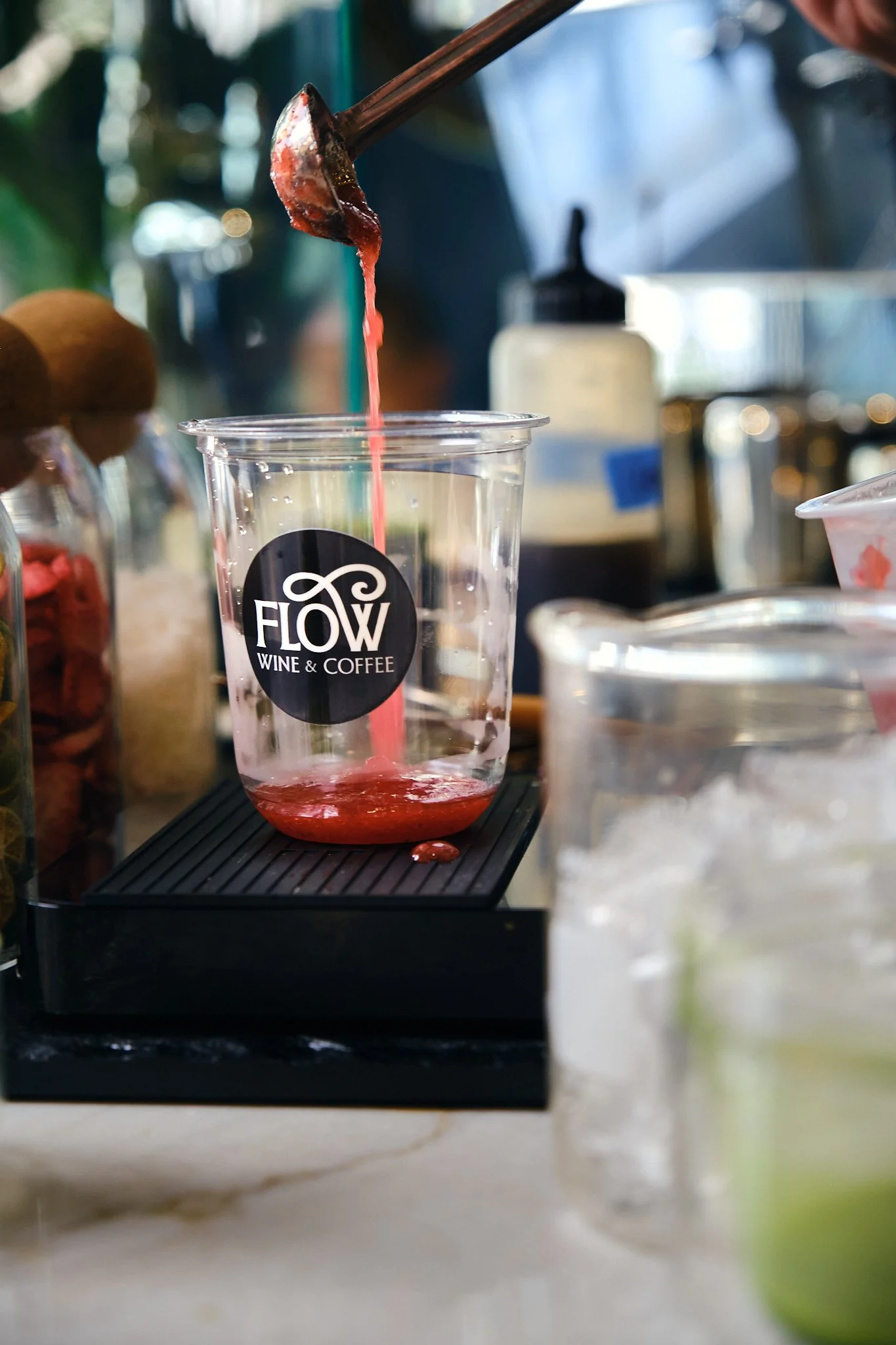 A clear plastic cup with the logo 'Flow Wine & Coffee' is on a black mat, with pink liquid being poured into it from a spoon. Various containers are blurred in the background.