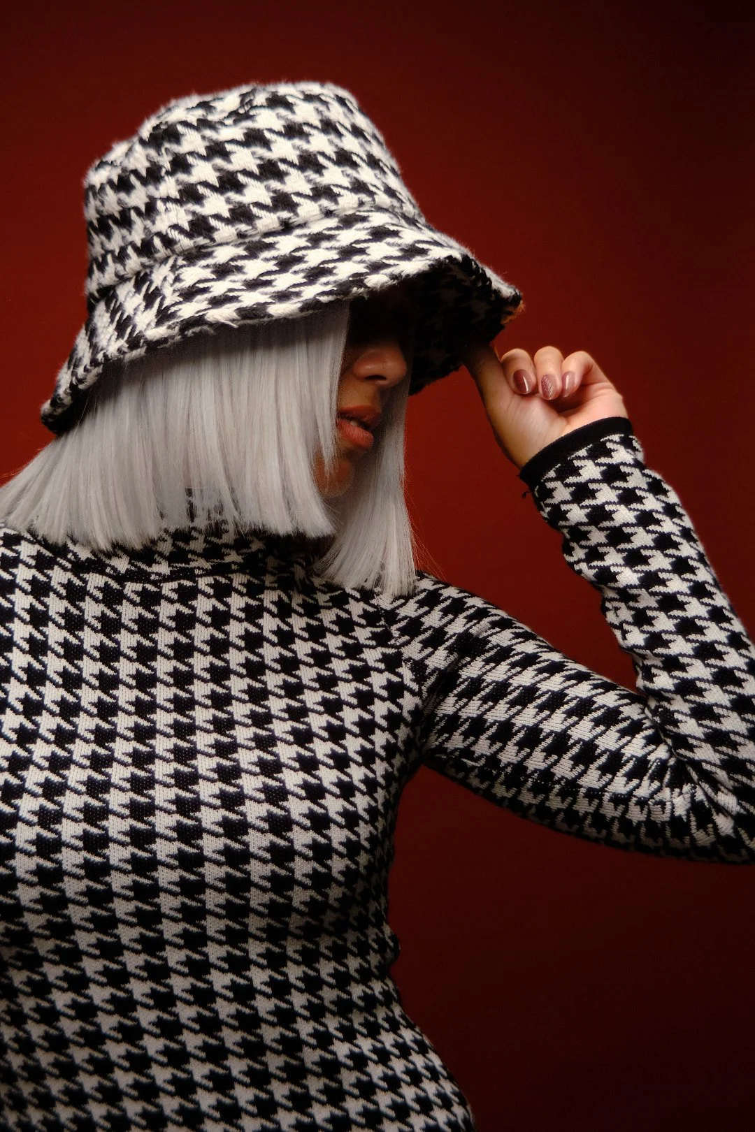 A woman with platinum blonde hair wearing a matching houndstooth pattern hat and top. She is touching the brim of her hat with her right hand. The background is red.