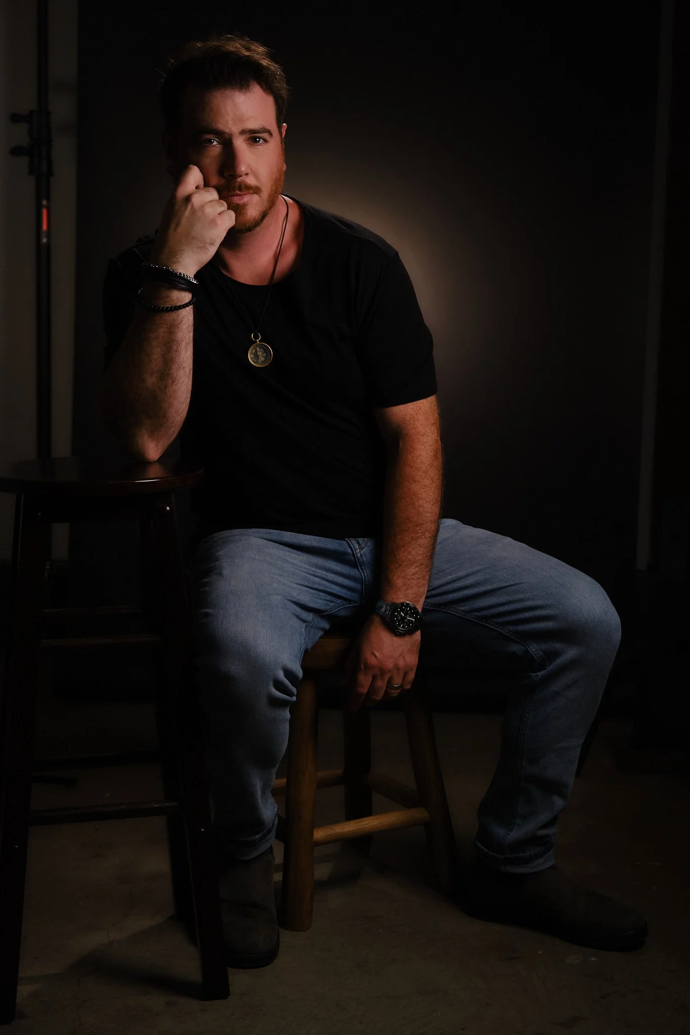 A man with a beard and dark hair sitting on a wooden stool, wearing a black t-shirt, light blue jeans, and black boots, with accessories including a wristwatch, bracelets, and a necklace with a pendant. He is resting his chin on his left hand and loo