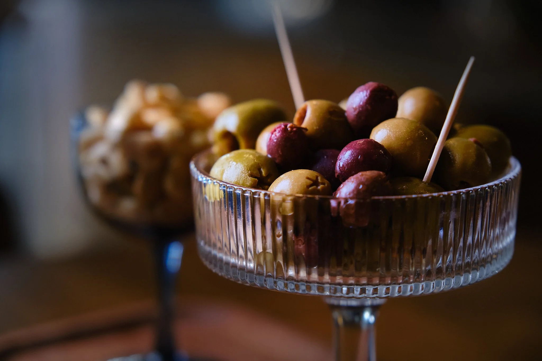 Close-up of a glass bowl filled with mixed green and purple olives, some with toothpicks inserted, set on a dark surface.