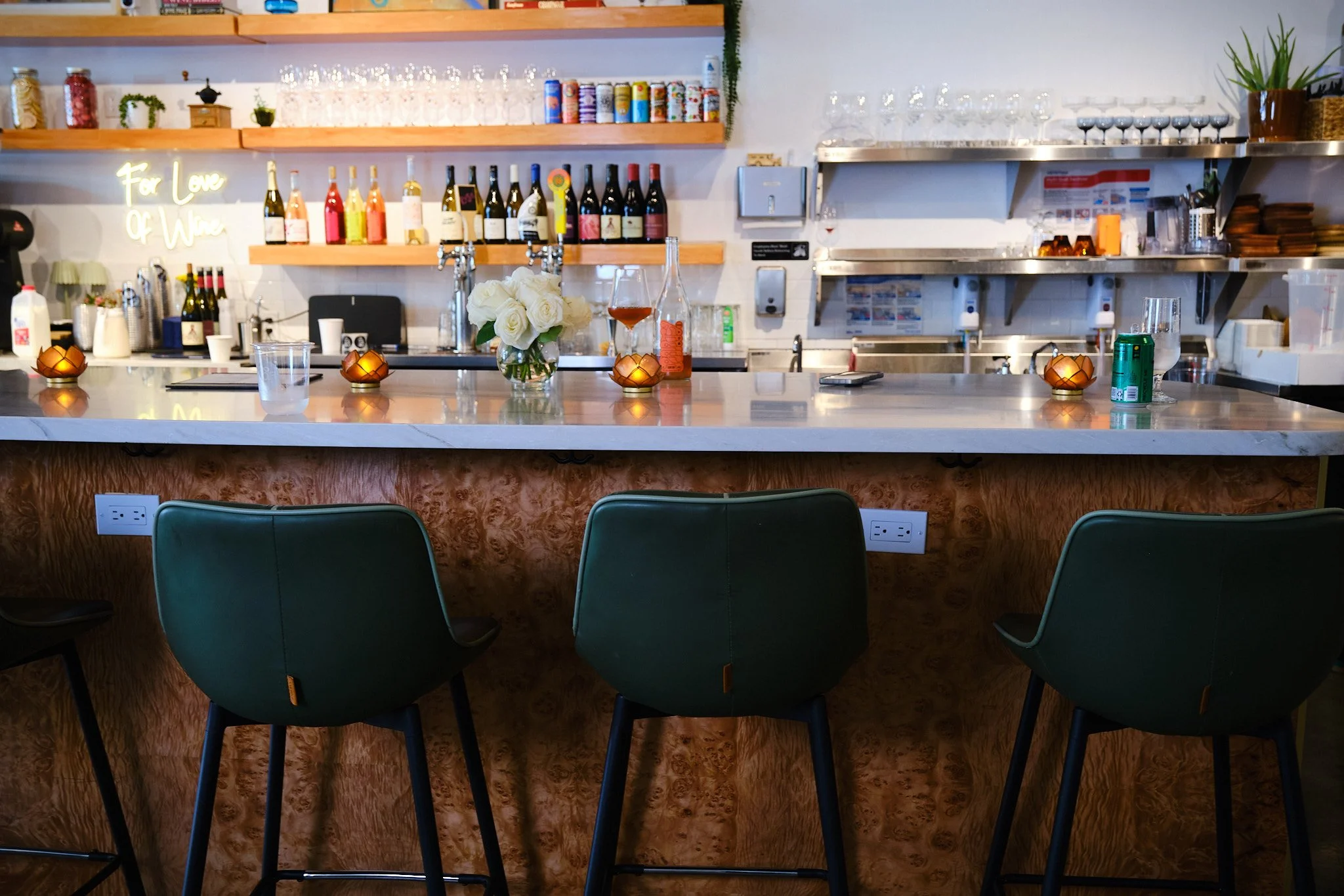 A bar or restaurant counter with three blue chairs in front, decorated with a vase of white roses and small candleholders. The back wall has shelves with wine bottles, glassware, and canned goods, and a neon sign that says 'For Love Of Wine.'