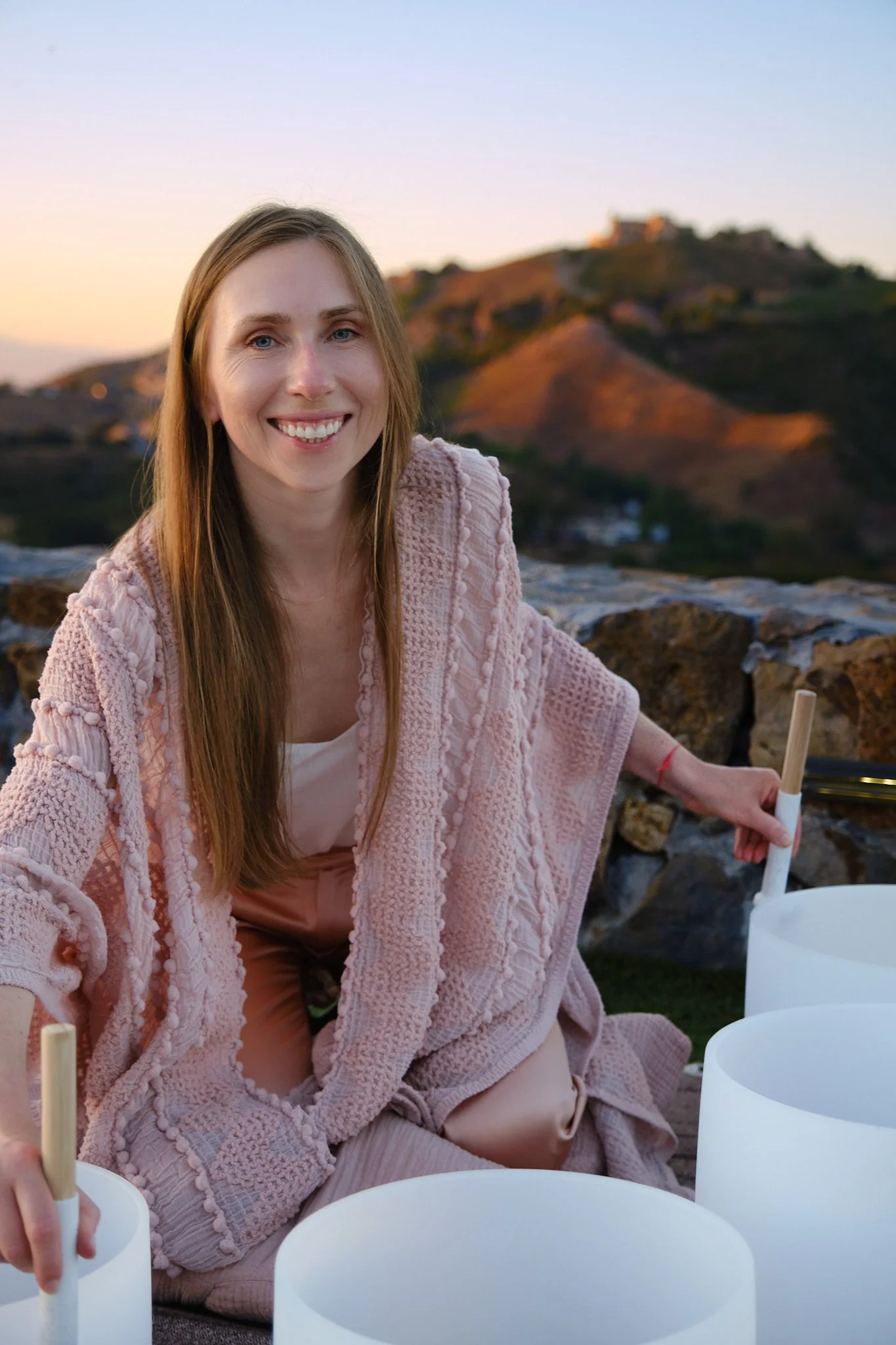 A young woman with long, blonde hair and blue eyes, smiling and wearing a pink textured coat, sits outdoors during sunset with a hill and buildings in the background, surrounded by white lanterns and holding a stick.