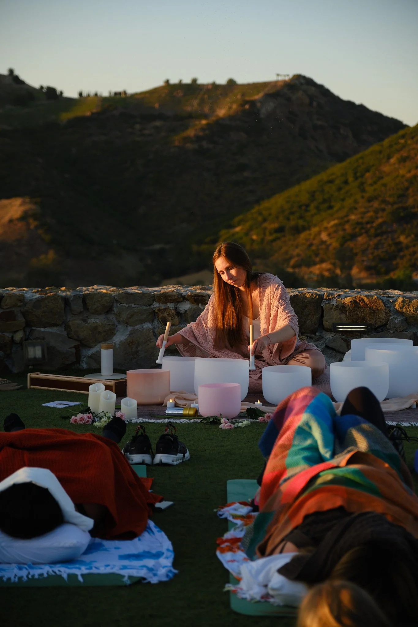 A woman in a pink knitted shawl or cardigan, sitting cross-legged on a blanket, is performing a musical activity with large white singing bowls outdoors. She is surrounded by various candles and flowers, with a stone wall behind her and mountainous terrain in the background during sunset.