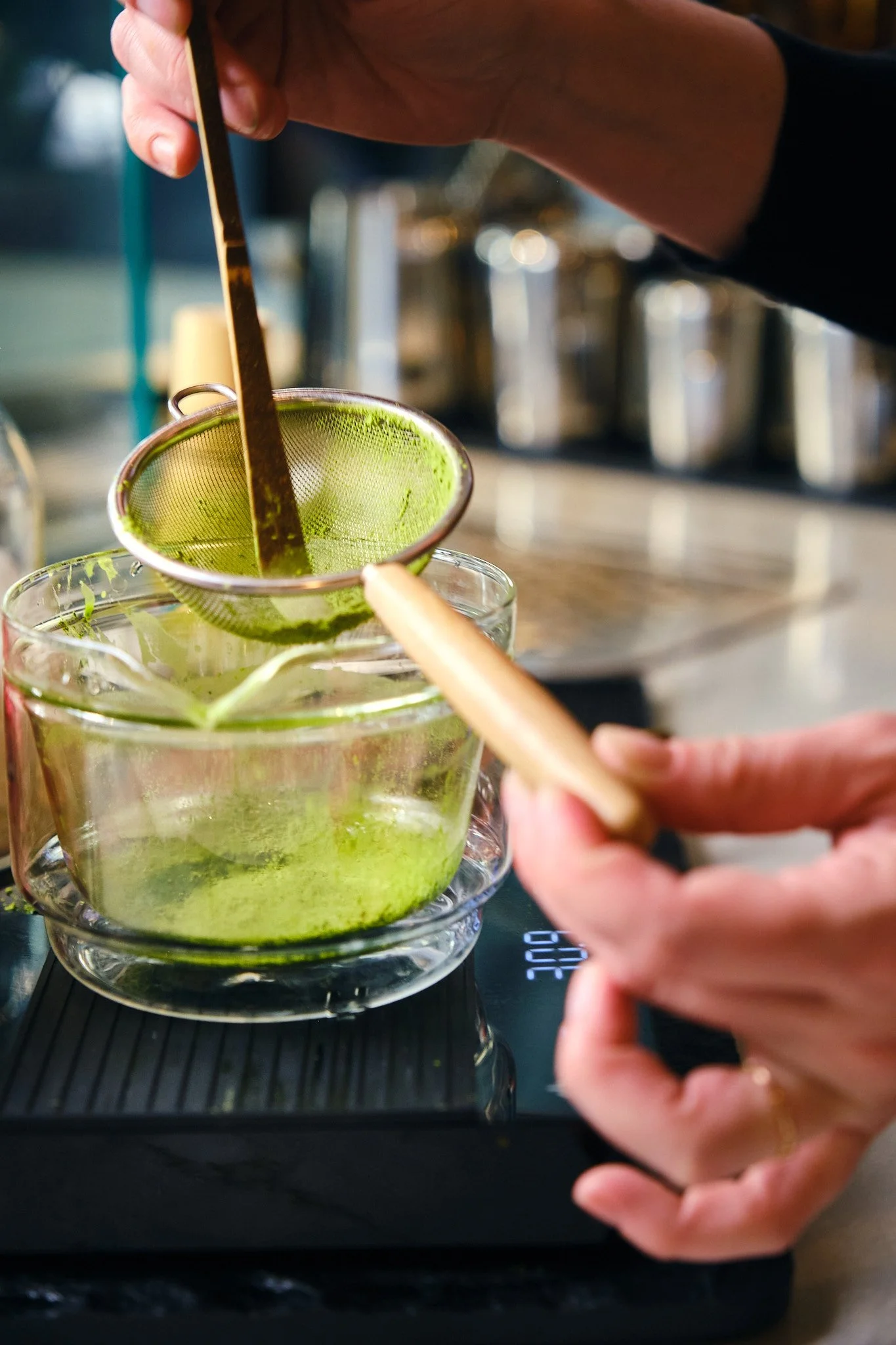 A person is using a tea infuser to steep green tea in a glass cup. The person's right hand holds the infuser, and their left hand holds a wooden stick, possibly to hold the infuser's handle. The background includes a kitchen setting with jars or cont