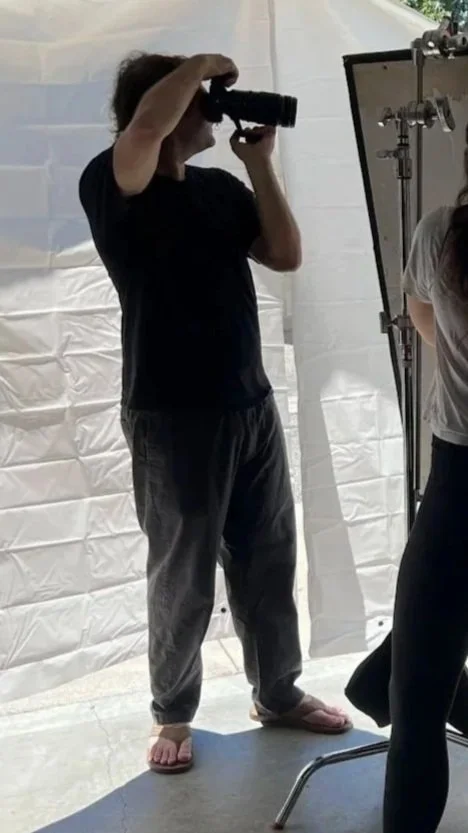 Person standing barefoot, looking through a camera or binoculars, inside a studio or photobooth setting with a white backdrop and equipment.