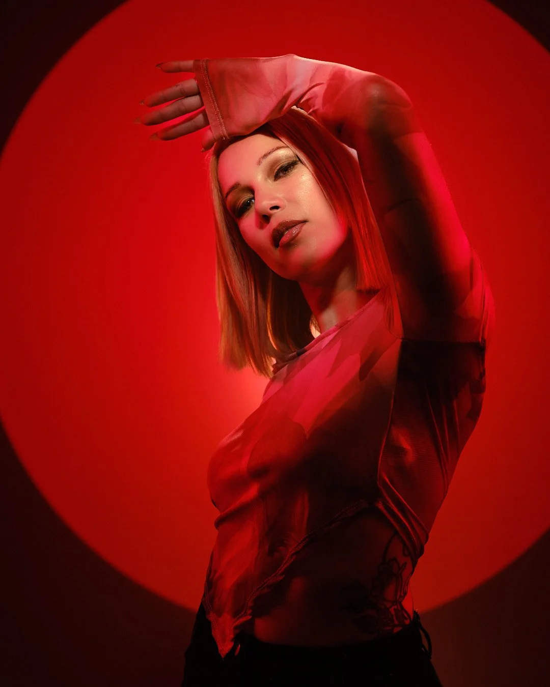 A woman with straight, shoulder-length hair posing with her arm raised and her hand resting on her forehead against a red circular backdrop.