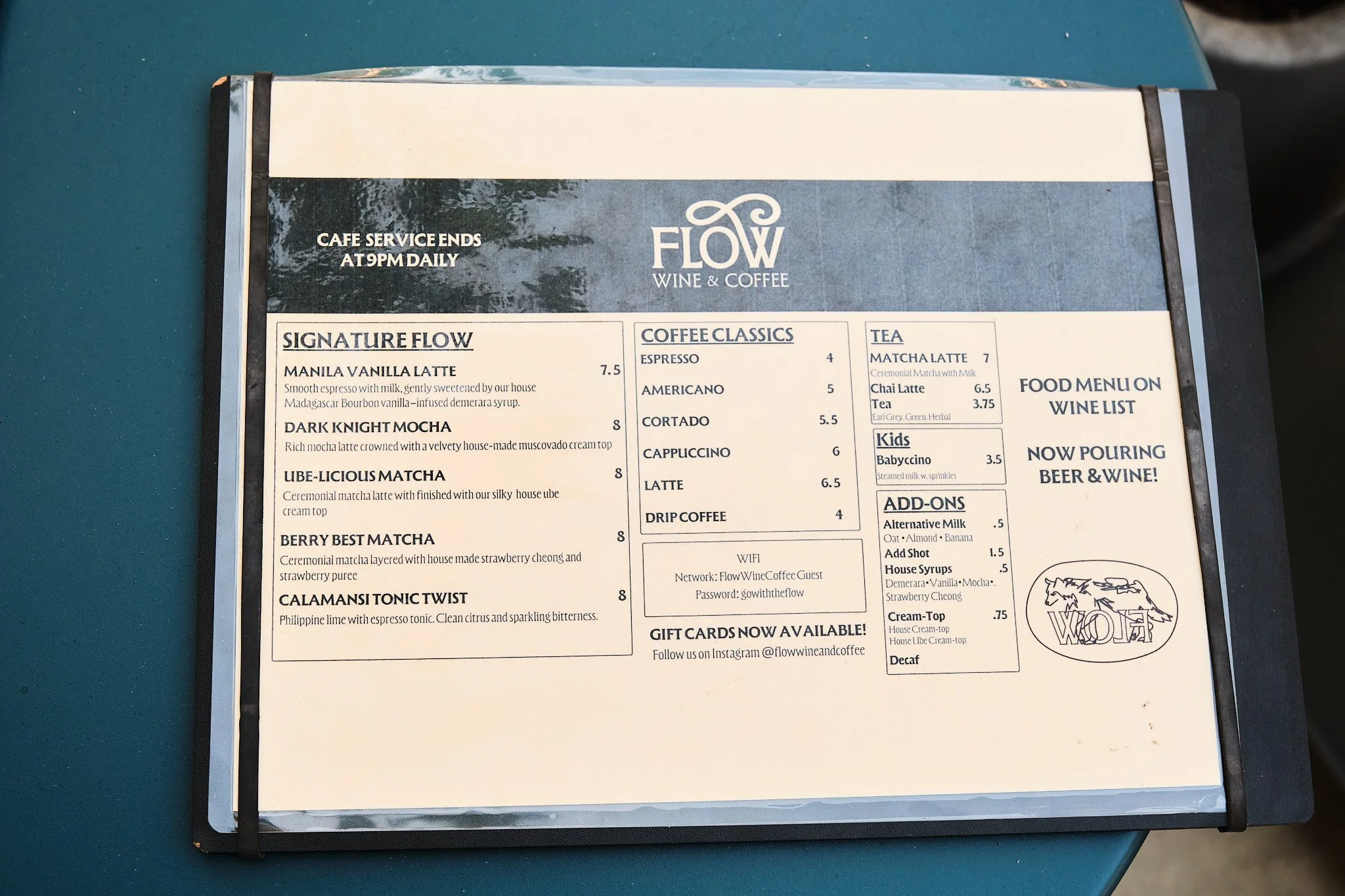 Printed menu from Flow Wine & Coffee, placed on a blue table. The menu includes sections for Signature Flow coffee, Coffee Classics, Tea, Kids, Add-ons, Food Menu, Wine List, and an announcement that they are now pouring beer & wine. It also mentions