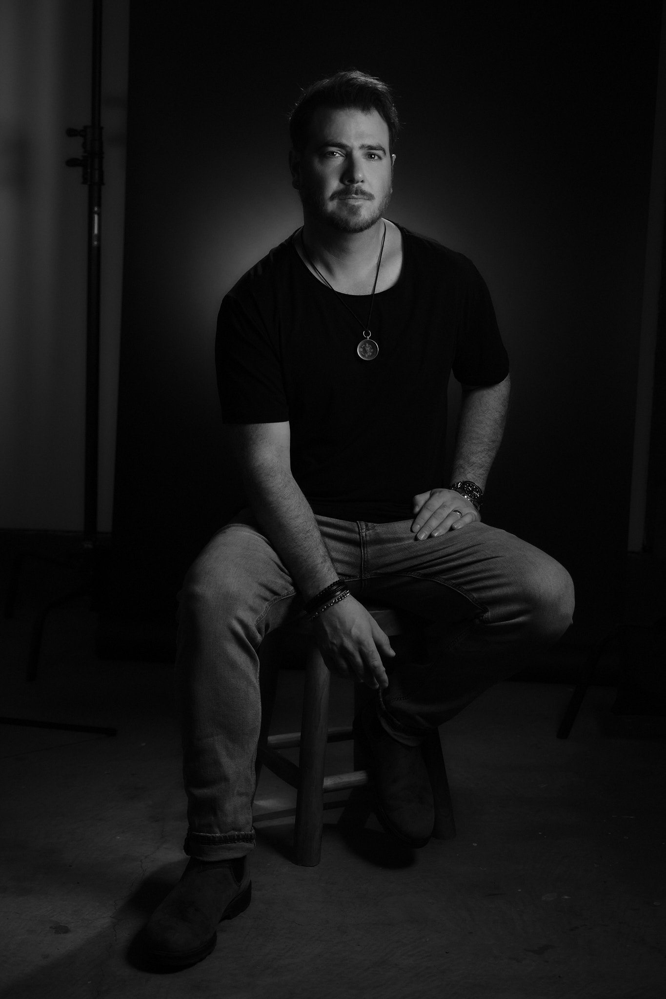 A black and white portrait of a young man sitting on a stool, wearing a black t-shirt, jeans, boots, and a necklace with a round pendant, in a studio with a dark background.