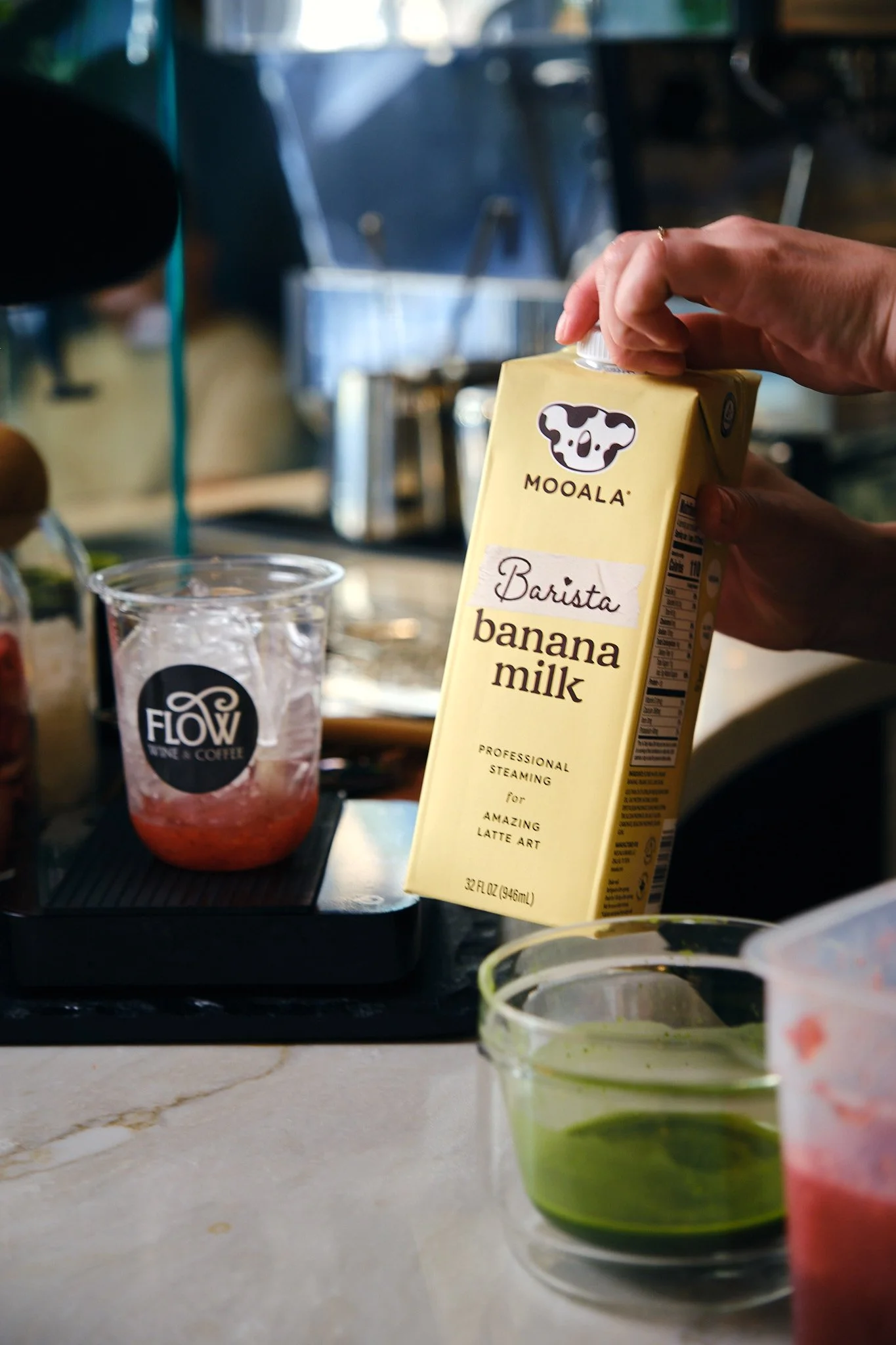 Hand pouring banana milk from a yellow carton with a cow logo into a clear glass on a countertop.