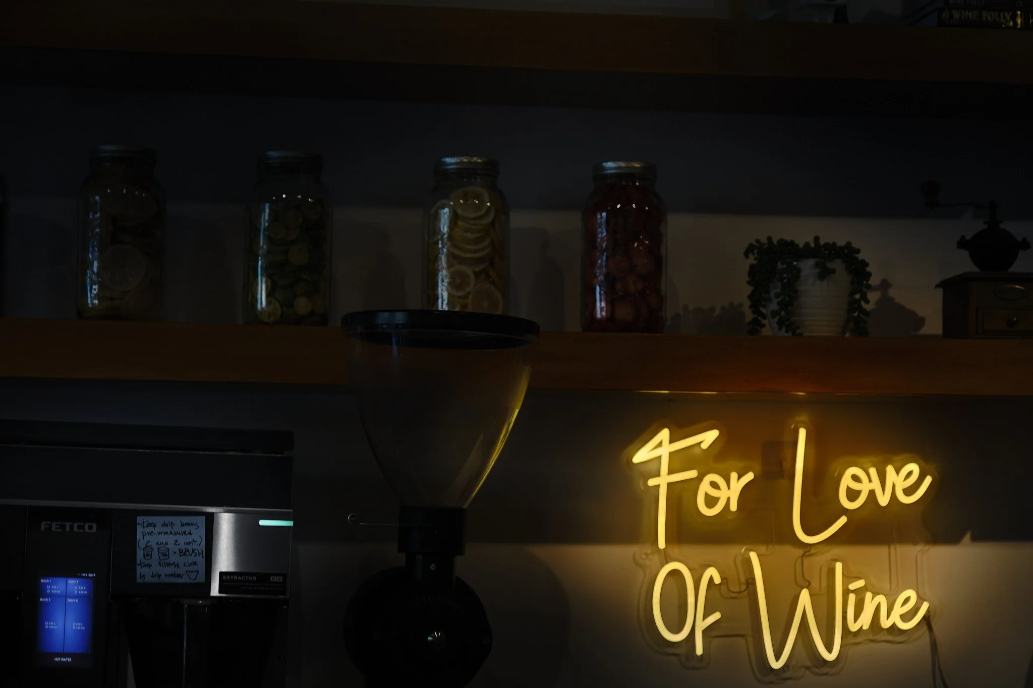 Jars filled with lemon slices, pickles, and red cherries on a shelf over a neon yellow sign that reads 'For Love of Wine' in a dimly lit setting.