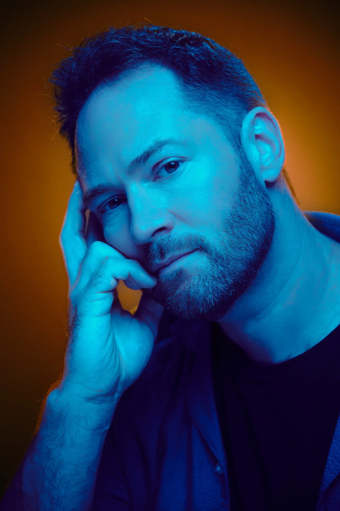 Close-up of a man with a beard looking at the camera, resting his face on his hand, with a brown background and blue lighting.