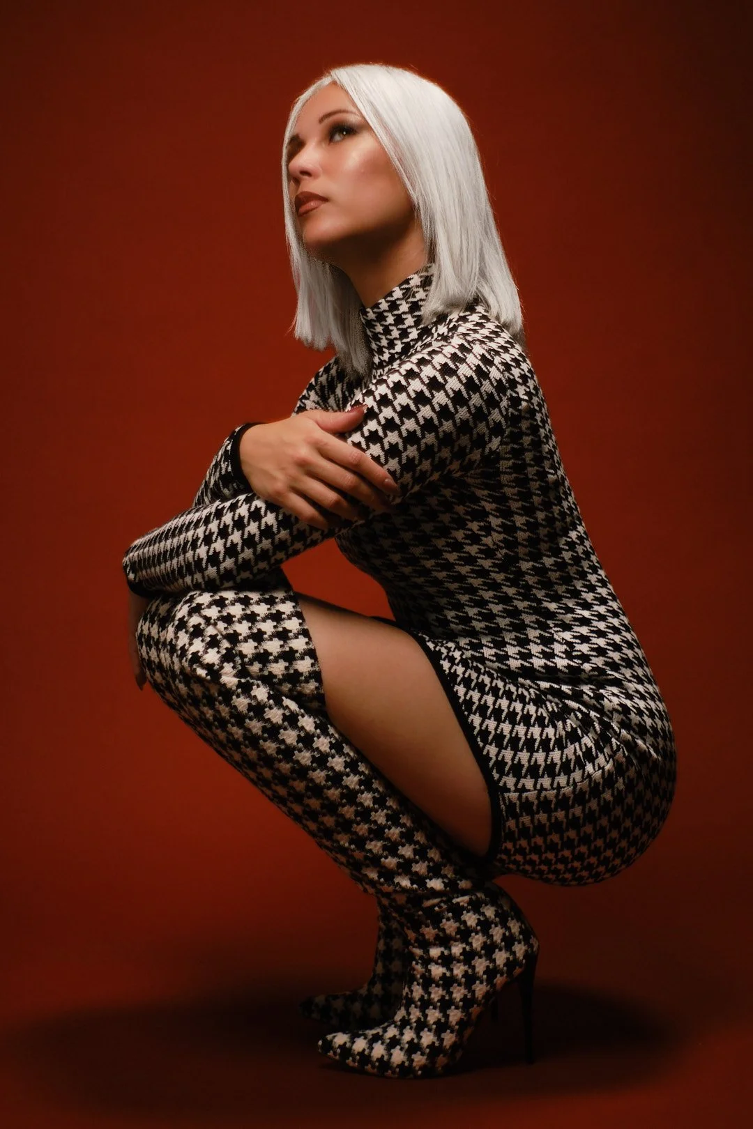 A woman with platinum blonde hair in a houndstooth patterned dress and matching thigh-high boots, squatting against a plain rust-colored background.