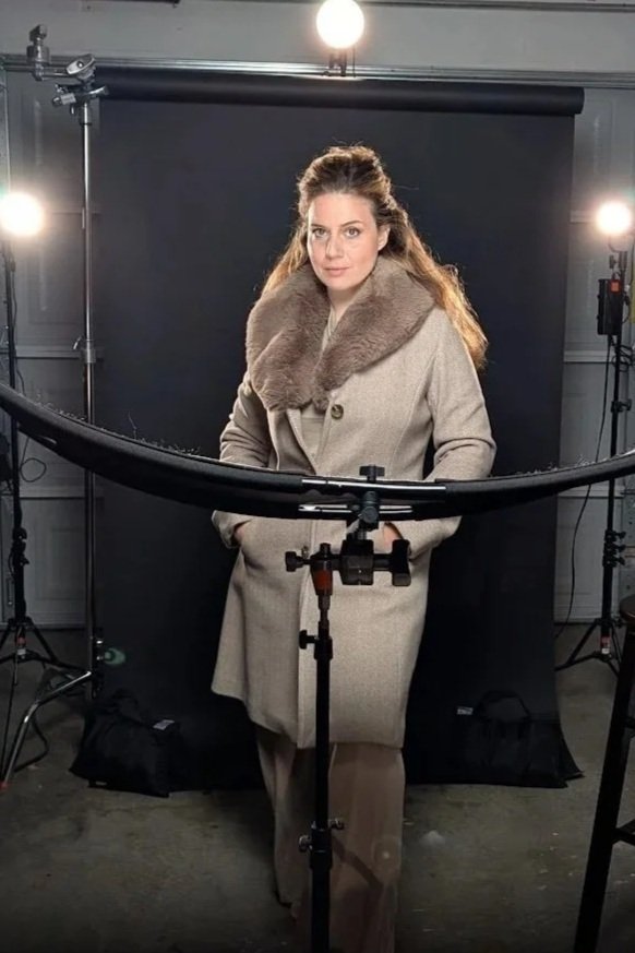 Woman standing in front of a black backdrop in a studio, illuminated by studio lights, wearing a beige coat with a fur collar, looking at the camera.