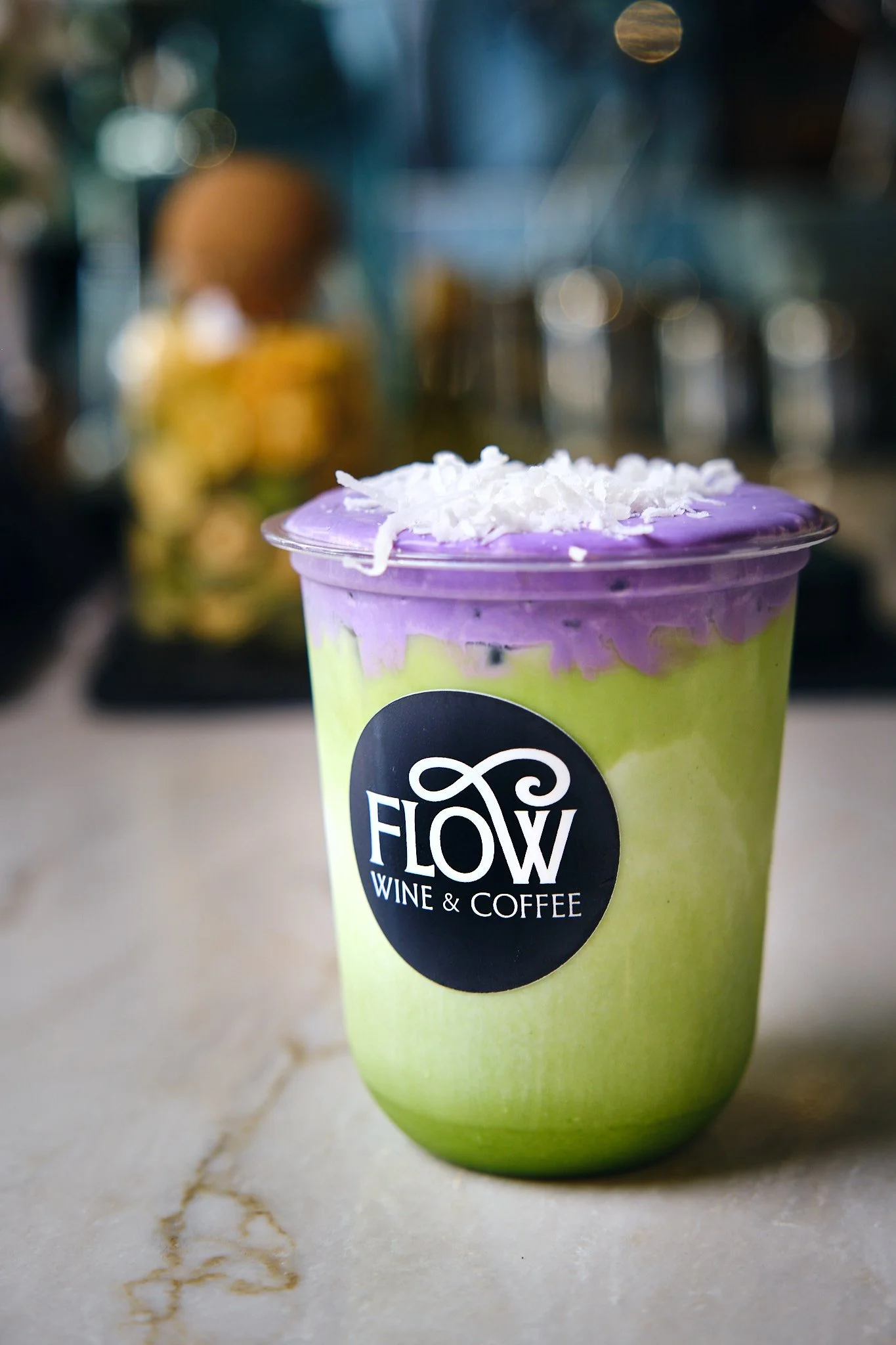 A colorful smoothie in a clear cup with a purple top and white shredded coconut on top, featuring a black circular label with the words 'Flow Wine & Coffee' on a light-colored marble surface.
