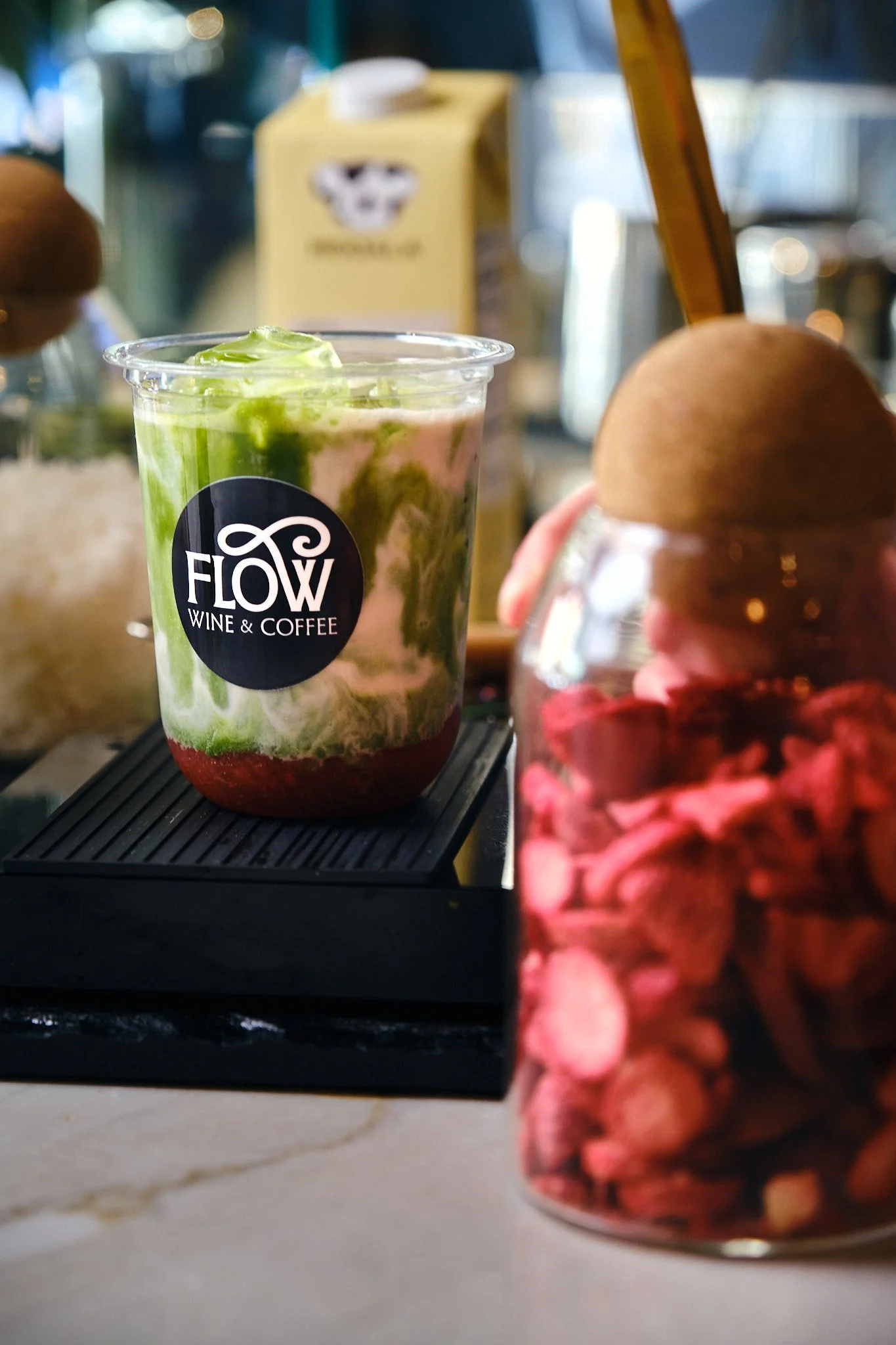 A clear plastic cup with a logo that reads "Flow Wine & Coffee," filled with a layered beverage consisting of red, white, and green sections, topped with whipped cream and green mint leaves. In the background, a blurred yellow napkin dispenser and ot