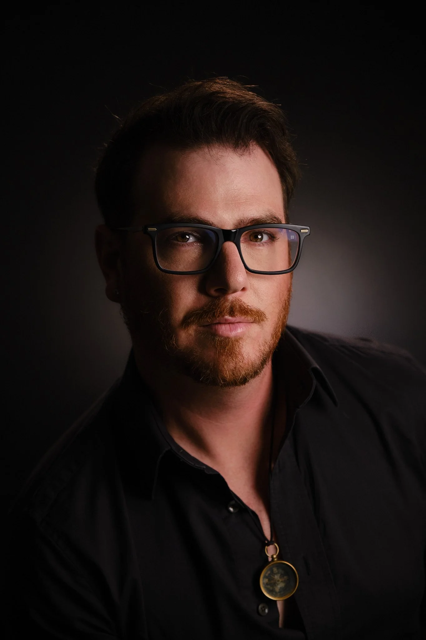 Portrait of a man with brown hair and beard wearing black glasses and a black shirt, against a dark background.