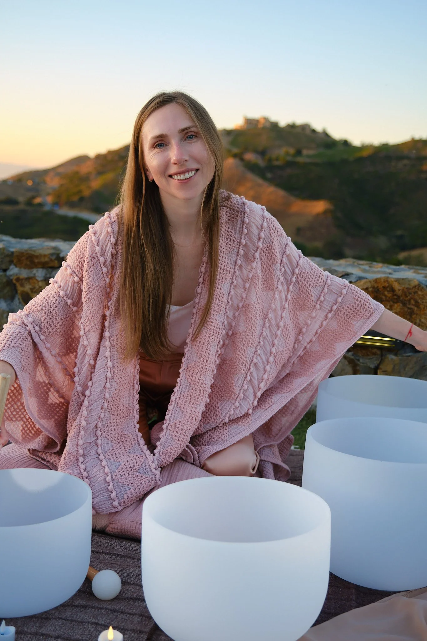 A woman with long hair smiling at the camera, wearing a pink textured shawl, outdoors with hills and houses in the background during sunset, surrounded by white decorative lanterns.