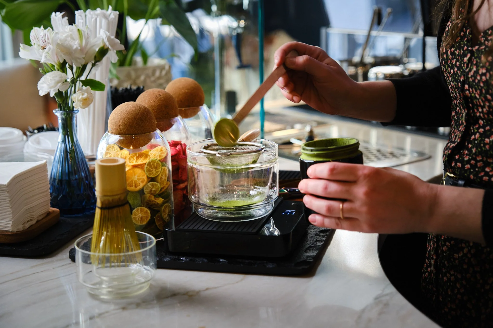 A person making a cocktail behind a bar or counter, with ingredients and flowers nearby.