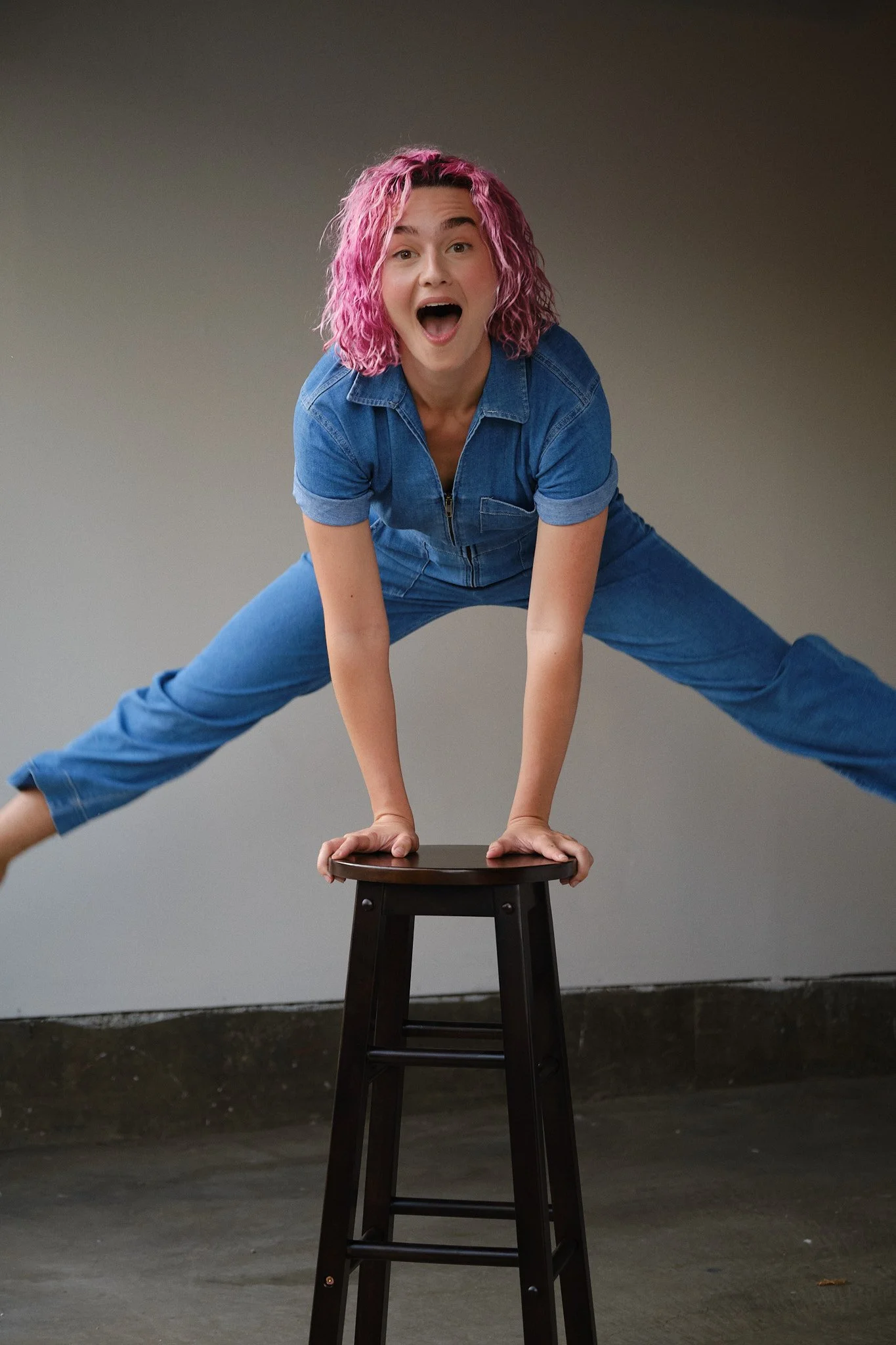 A woman with pink curly hair in a blue jumpsuit performing a handstand on a stool, with a surprised expression.