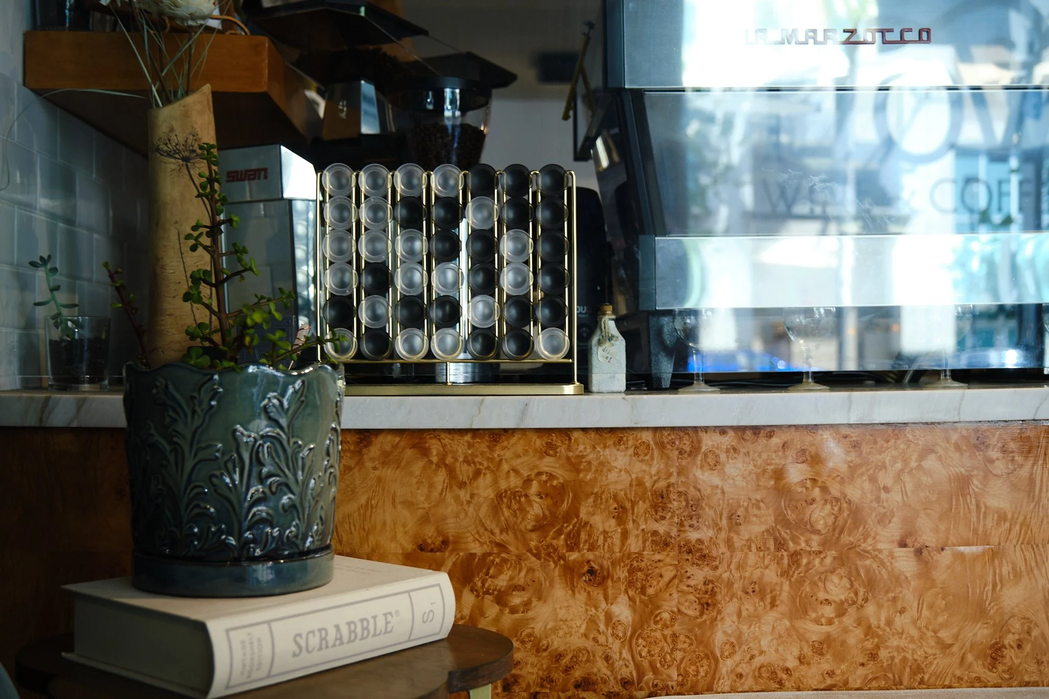 A display shelf with a plant in a decorative blue-green ceramic pot with embossed patterns, a box labeled 'SCRABBLE' on a stand in front of the shelf, and a coffee machine with coffee beans and containers around it in a cafe or coffee shop setting.